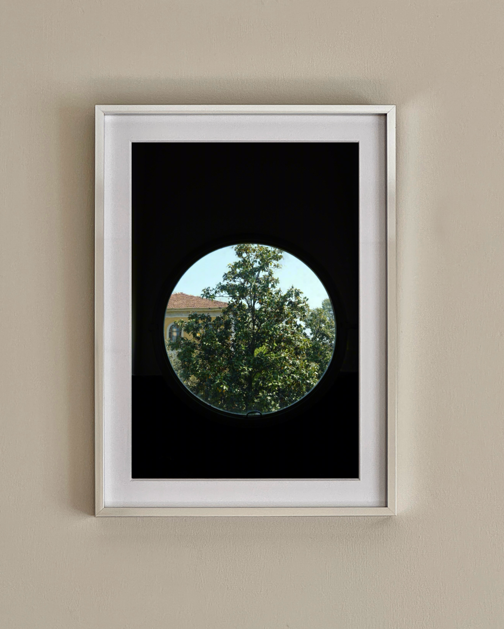 A framed limited edition "Villa Necchi Campiglio, Milan - Print Photography" by Grace Rose Watts hangs on a beige wall, depicting a tree and building through a circular window with a black background.