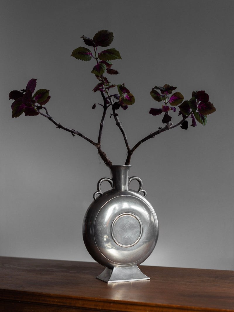 The round Spigel Art Deco Pewter Vase features a circular design and holds tall, bare branches adorned with dark purple and green leaves. This collectible vintage décor piece is displayed on a wooden surface against a plain gray background.