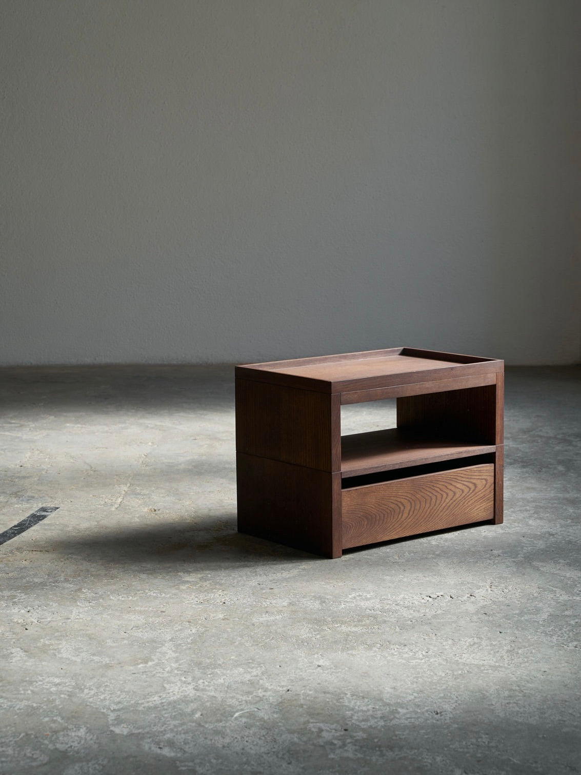 The Eileen Bedside Table + Tray by ASERIES OF OBSTACLES, SL, is a minimalist wooden side table with a rich brown finish. It has a rectangular shape, an open shelf beneath the tabletop for multifunctional use on a concrete floor against a plain wall, casting shadows on the floor.