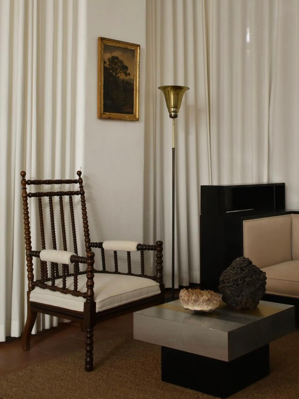 A cozy living room corner with a 19th-century charm showcases a pair of wooden armchairs by Barracuda Interiors, complemented by a tall brass floor lamp, a modern sofa, and a square coffee table adorned with decorative stones. White curtains and a framed painting add an elegant touch to this delightful English-inspired space.