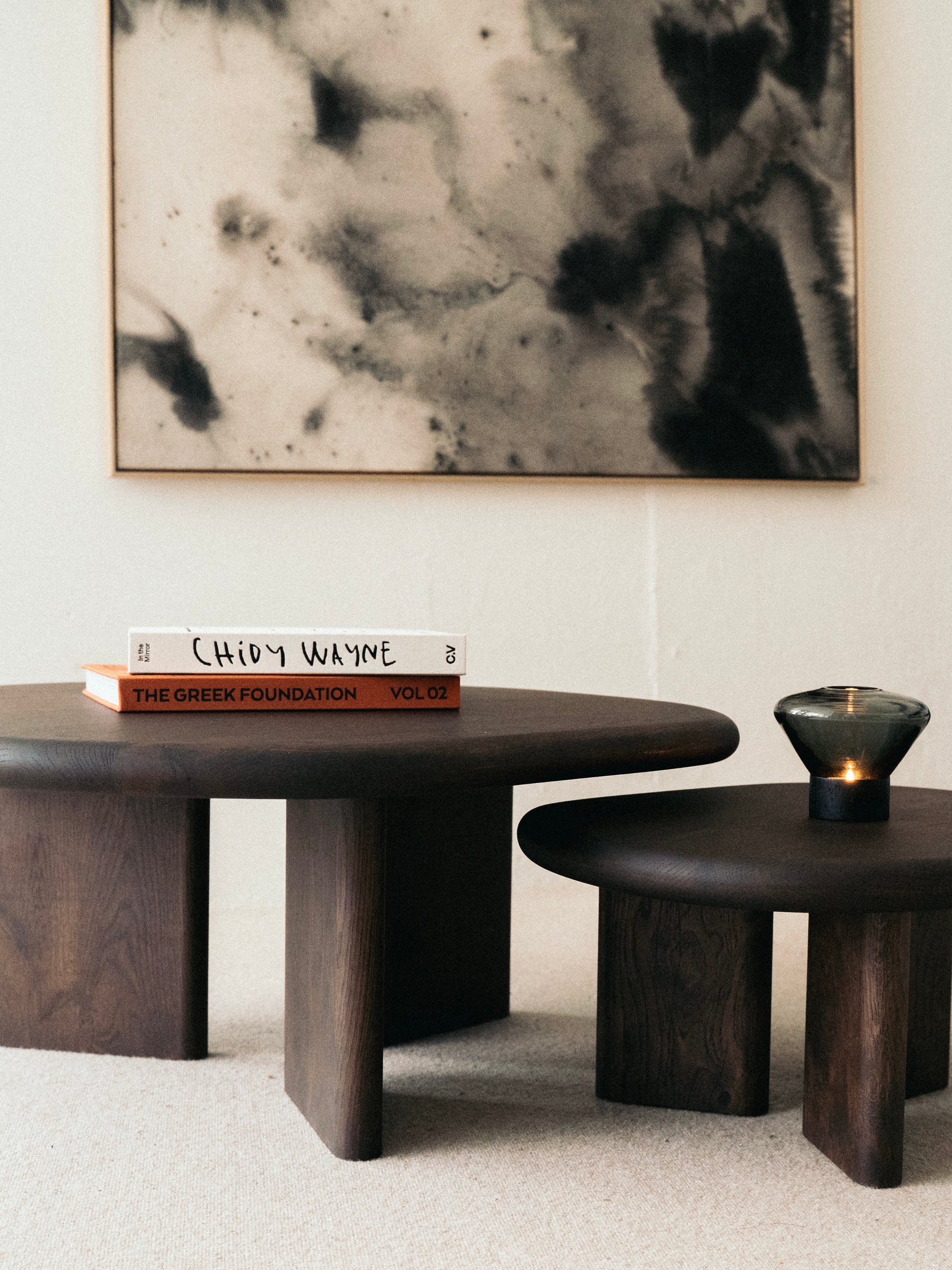 A modern living room setup featuring two dark wood tables, one larger and one smaller. The larger table, the KØGE Design Double Espresso Coffee Table, boasts a solid oak construction with an espresso-tinted varnish finish and is adorned with two books and a small, dark glass candle holder. A piece of black and white abstract art hangs on the light-colored wall in the background.