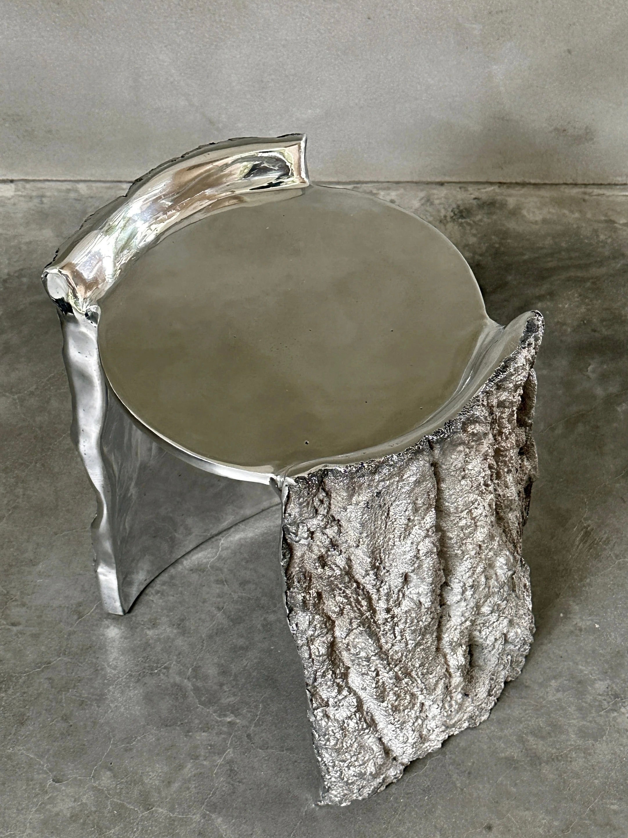 The "Bark" Side Table by Kaytar showcases a high-gloss metallic, reflective top with an Ironbark texture reminiscent of rough stone. Set on a concrete floor against a plain wall, it combines modern elegance and rustic charm.