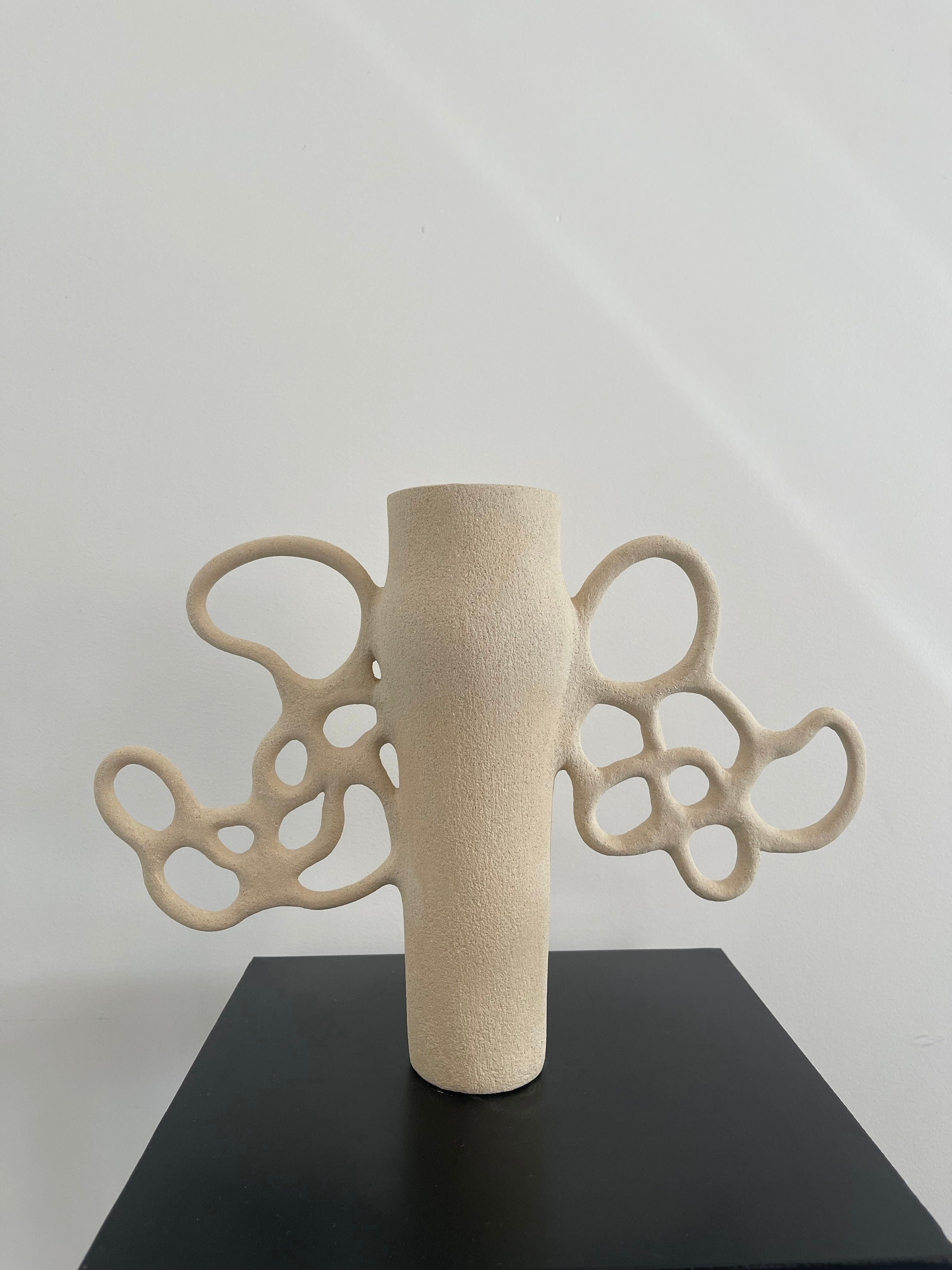 A beige, textured Vase Liens n°1 from Louana Roudot Ceramic stands on a black pedestal, its wavy, abstract handles resembling intricate organic loops crafted in white chamotte stoneware. The background is plain and light-colored.