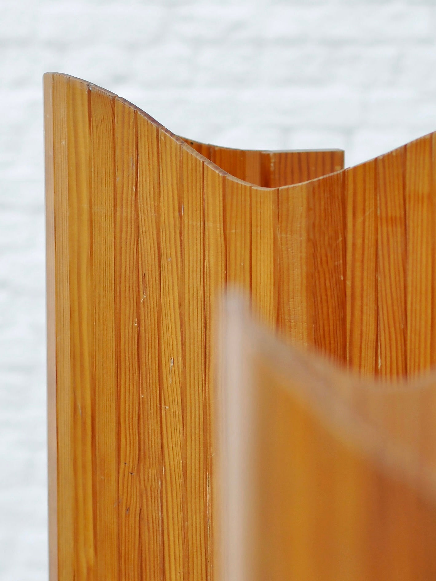 Close-up of the Jomaine Baumann Room Divider by Introverso, 1940s. It features a light brown wooden panel with a wave-like top edge against a blurred white brick wall. The tambour design highlights the wood grain, exuding the elegance and natural texture synonymous with French Art Deco style.