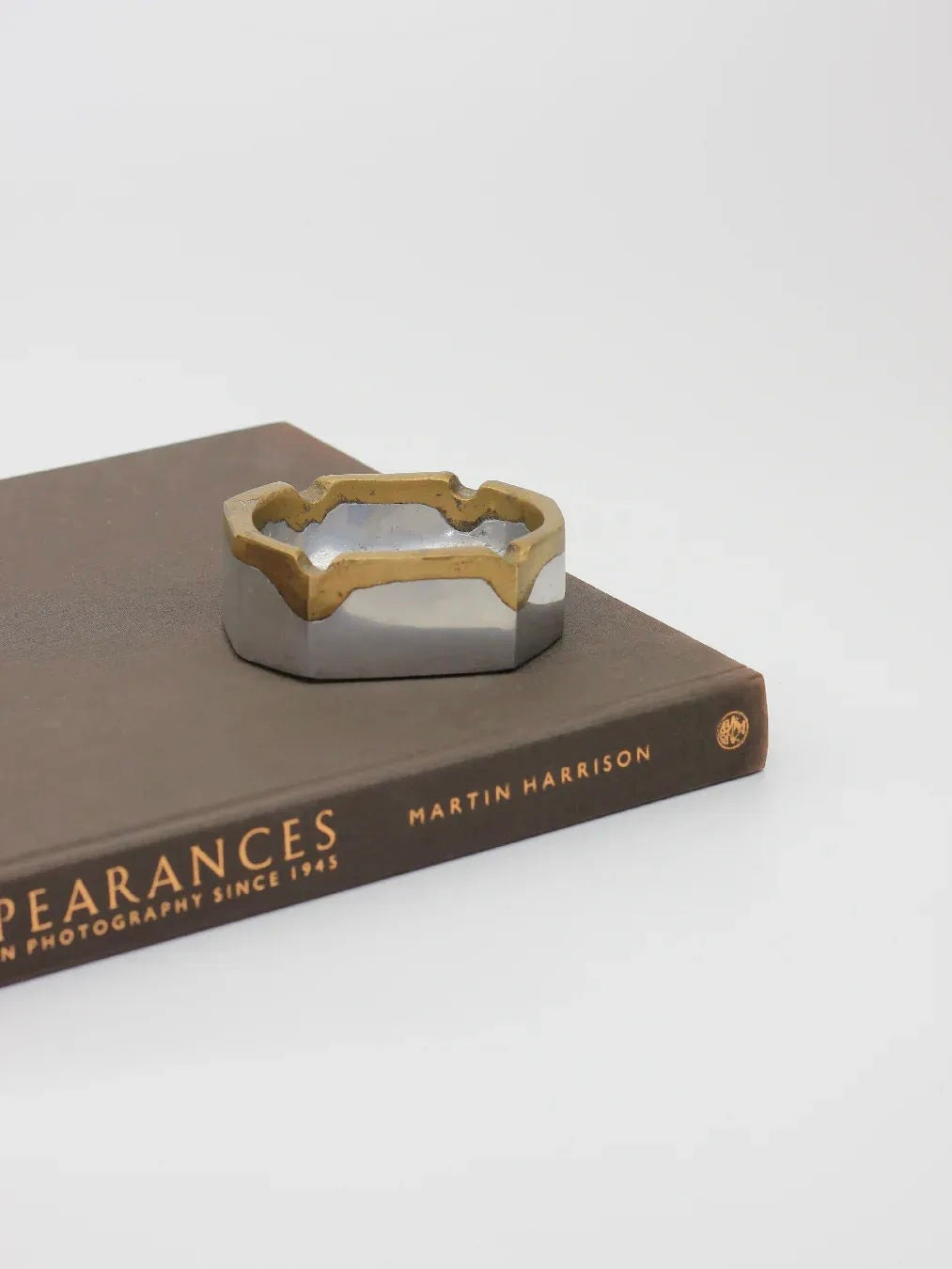 A Brutalist ashtray from Boga Avante Shop, featuring a silver and gold finish with an irregular edge, is placed atop a brown book titled "Appearances" by Martin Harrison. Its bold design evokes David Marshall's aesthetic against a plain white backdrop.