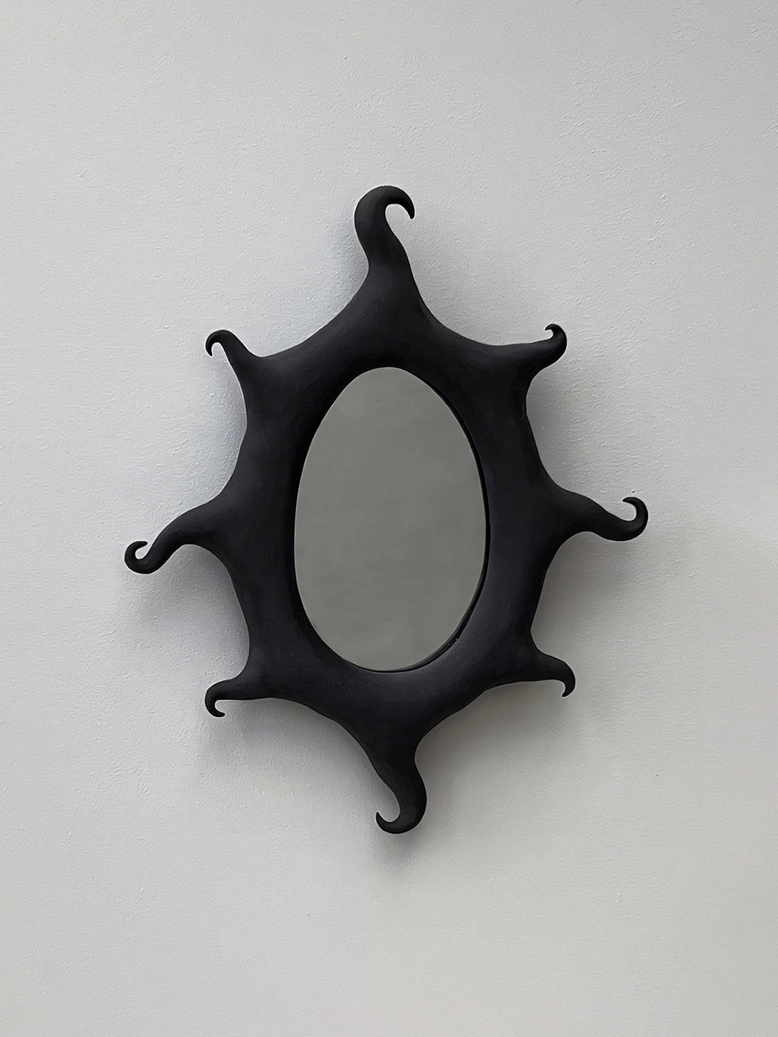 The Black Sun Mirror by Solenne Belloir, a handcrafted ceramic piece featuring an oval shape and a distinctive tentacle-like frame in a black matte finish, is mounted on a plain white wall, exuding a magical presence.