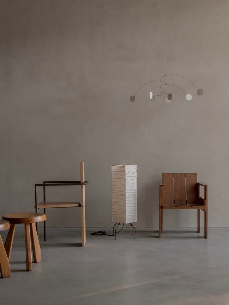 The minimalist room with pale walls includes a wooden stool, a simple chair, a narrow table, and an elegant vintage Akari UF lamp by Bicci de' Medici on a black stand. A modern mobile hangs from the ceiling, epitomizing mid-century modern style.