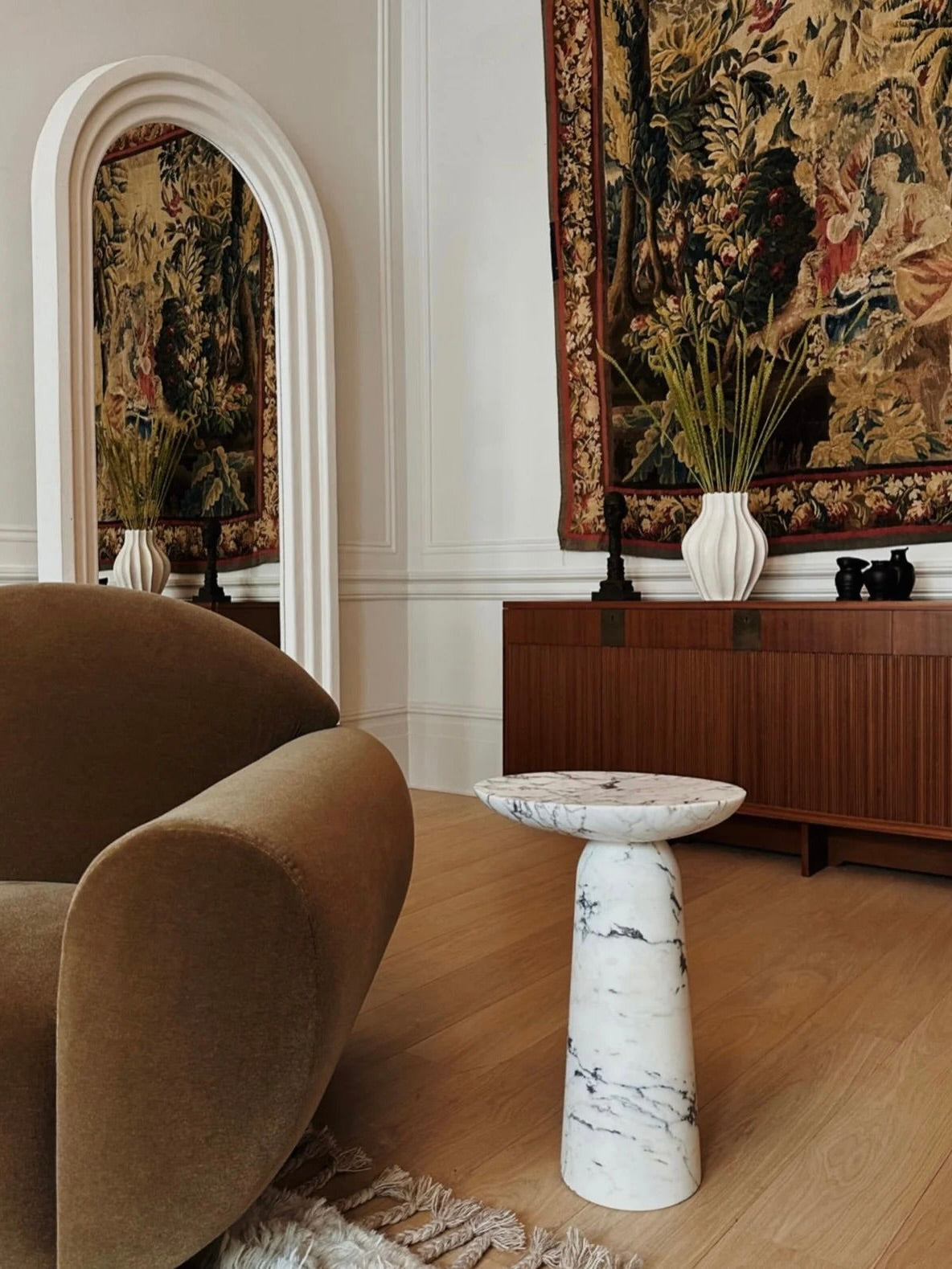 A stylish living room showcases a curved brown chair, a Ransom & Dunn Athena Marble Side Table with white Calacatta Viola marble, and a wooden cabinet. A large ornate tapestry is displayed above the cabinet, while a tall mirror with an arched frame stands beside it. A fringed rug adorns the wooden floor.