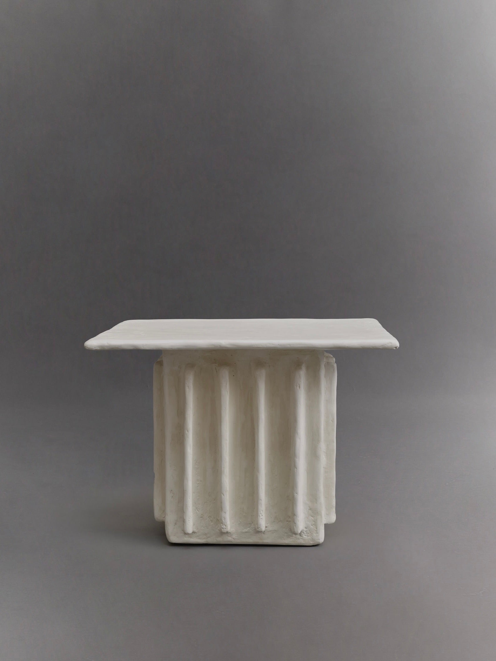 The Columnar Side Table by Ombia Studio is a rectangular, white ceramic table featuring a raw white plaster base inspired by sculpted columns, set against a plain gray background.