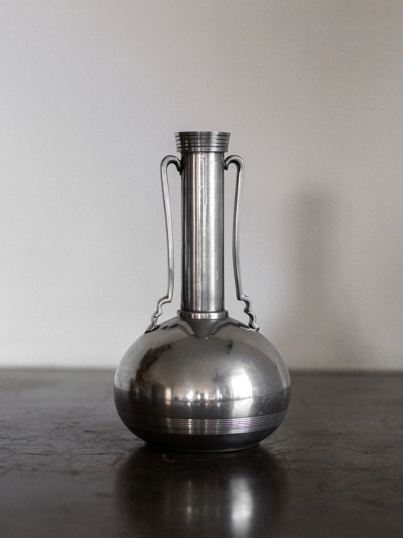 A polished silver Art Deco Metal Vase from 1935 by Spigel, featuring a rounded base and a long, slender neck with two thin, ornate handles, stands on a plain dark surface against a neutral background. Emblematic of Scandinavian craftsmanship, this vase boasts a minimalist yet elegant design.