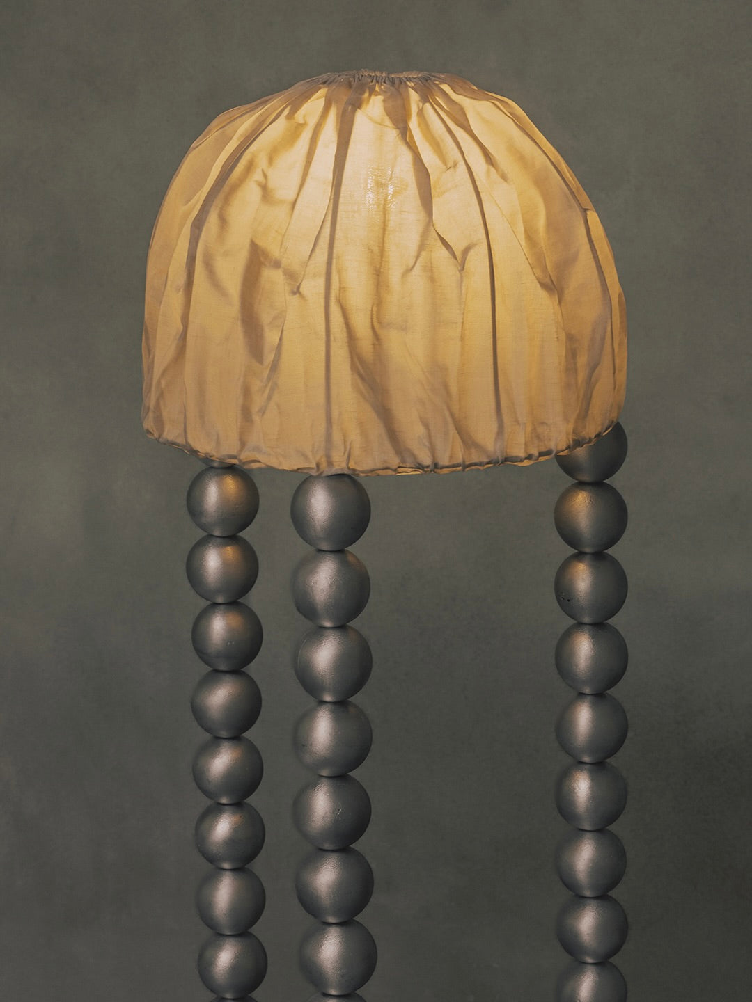 Introducing the "ALTER" Floor Light by Kaytar, featuring a handcrafted design with a soft beige linen shade resembling a jellyfish and supported by an alchemy-inspired trio of metallic aluminum spheres against a muted dark green backdrop.