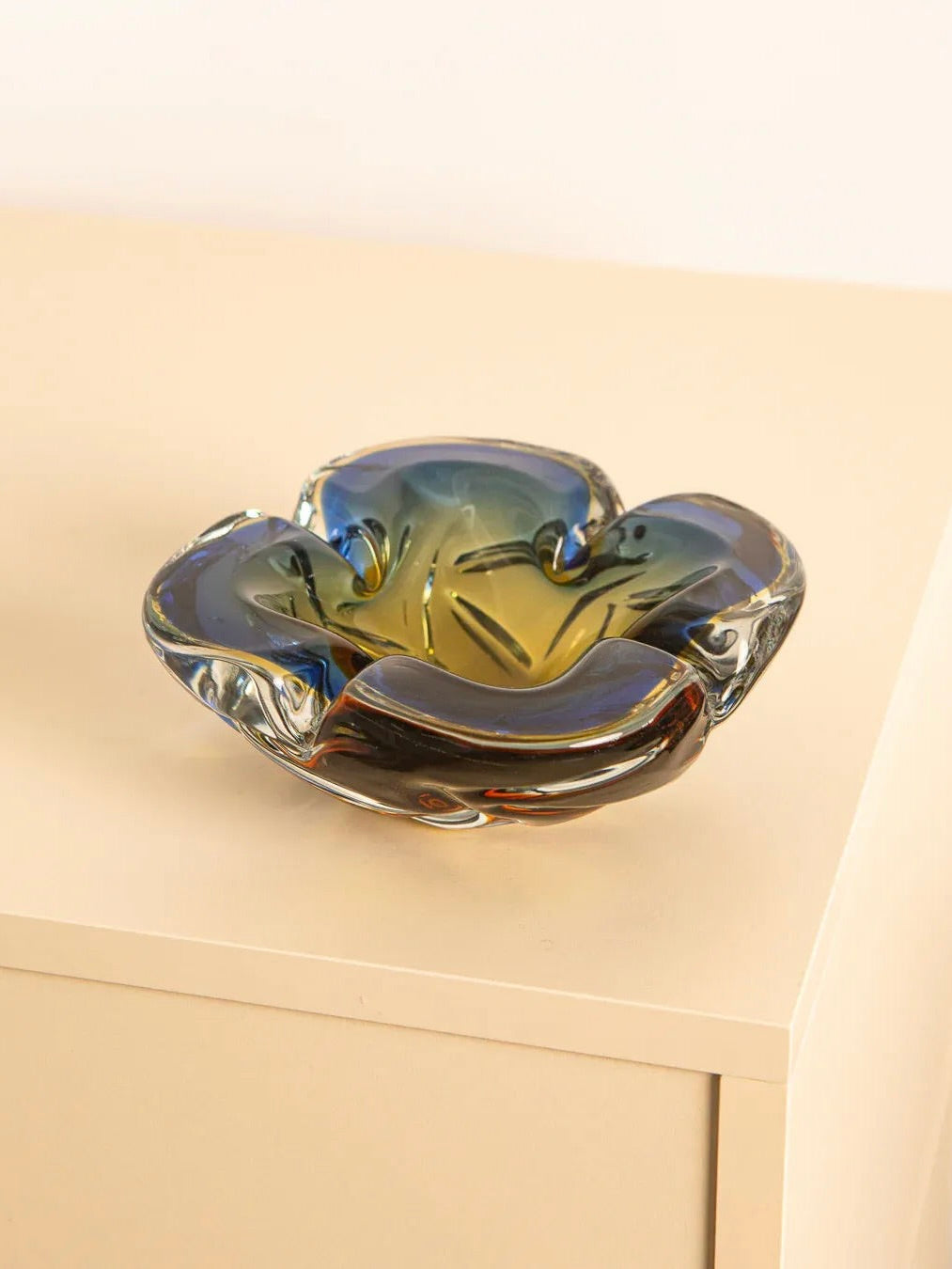 The Large Murano Glass ‘Fleur’ Vide Poche 60's by Treaptyque sits on a light surface. Its wavy design and gradient shades of blue, amber, and green reflect light with a smooth, glossy texture, showcasing exquisite Italian craftsmanship.