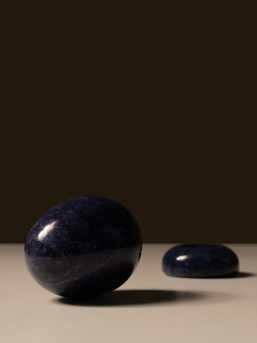 A close-up of The Marble Egg Sculpture by Les Objets Amsterdam, featuring two dark, polished stones on a smooth, light-colored surface. The larger, rounder stone in the foreground resembles a Lapis marble egg, while the smaller, flatter stone is positioned farther back. A plain dark background creates a contrasting backdrop.