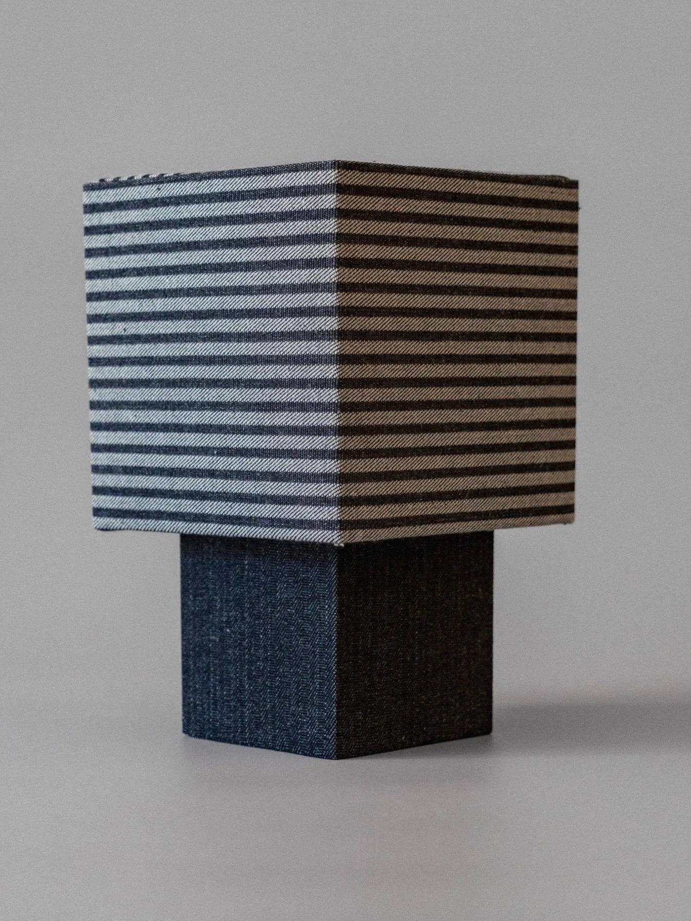 Introducing "La Petite Boîte," a contemporary lamp by mattina moderna featuring a gray and black striped cube-shaped shade crafted from deadstock denim. It is set upon a sleek, solid black rectangular base, embodying eco-friendly design against a simple, muted gray backdrop.