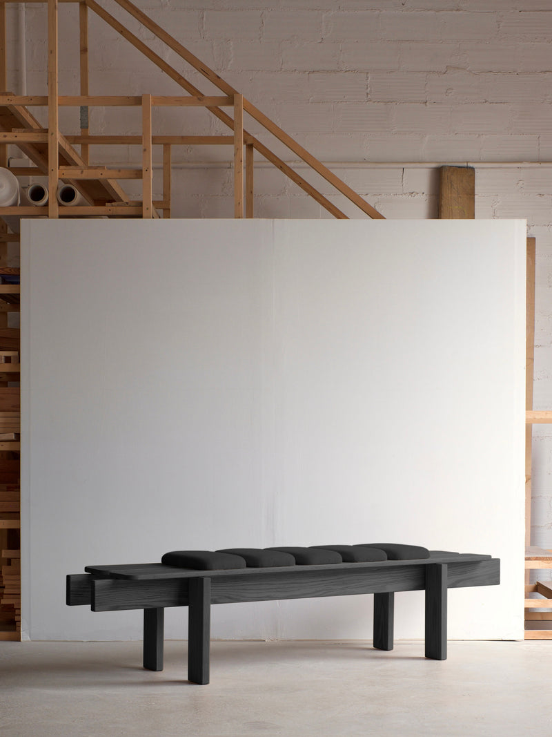 The Ueno Bench Black Edition by malva, with its modern tufted cushion, stands in a minimalist space, highlighting Japanese craftsmanship alongside a solid oak staircase and large white panel against a brick wall for a clean, industrial aesthetic.