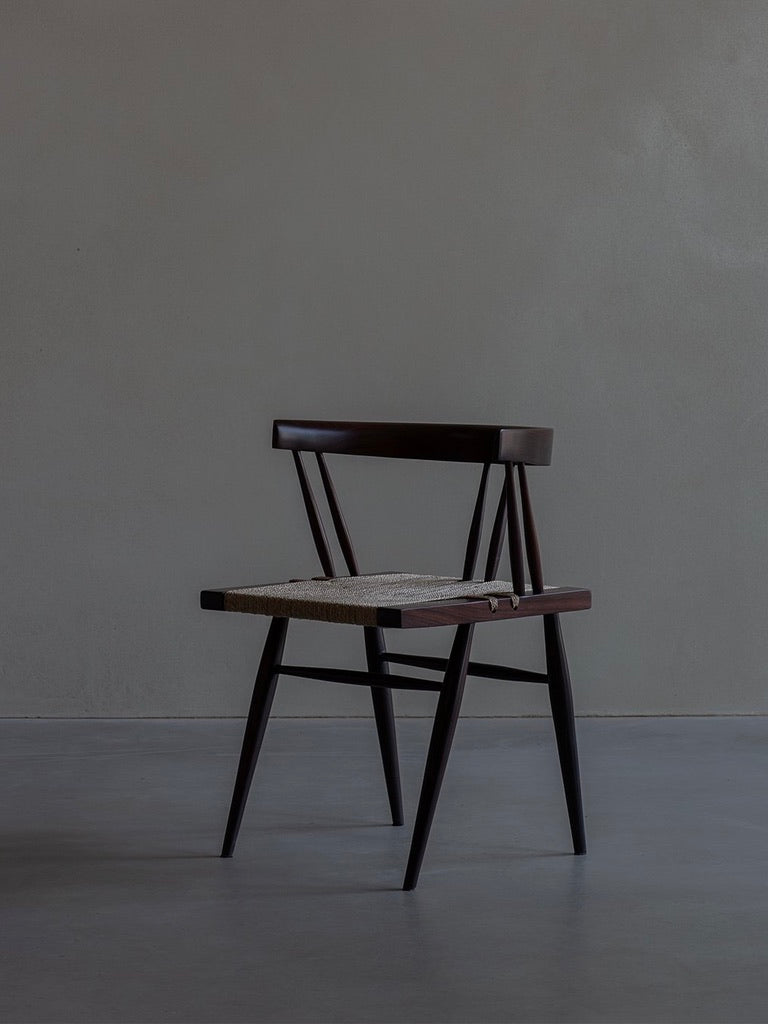 Against a plain gray wall stands the "Manner of Nakashima - Grass Seated Chair" by Bicci de' Medici. Crafted from dark rosewood, it features a horizontal backrest and woven seat. Its minimalist, mid-century style is beautifully accentuated by soft lighting.