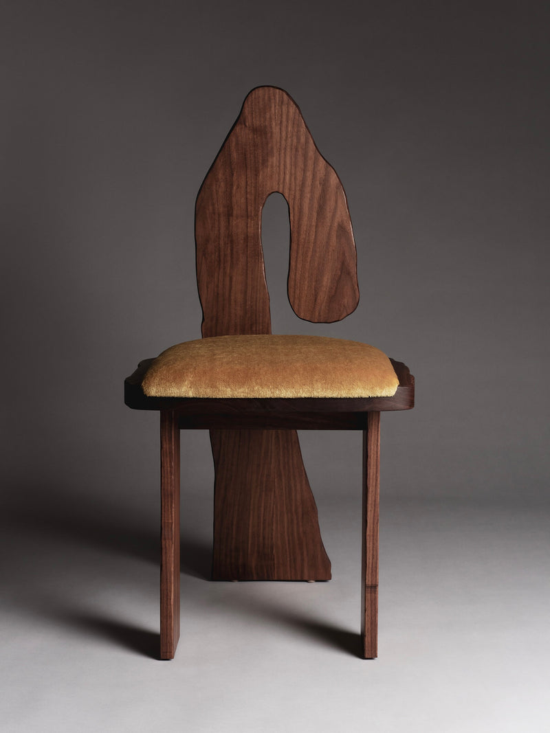 The Muisca Dining Chair by Ombia Studio is a distinct piece with a wavy, abstract wooden backrest and a mustard mohair-upholstered seat. Its solid oiled walnut frame features four rectangular legs, offering an elegant contrast against a neutral background.