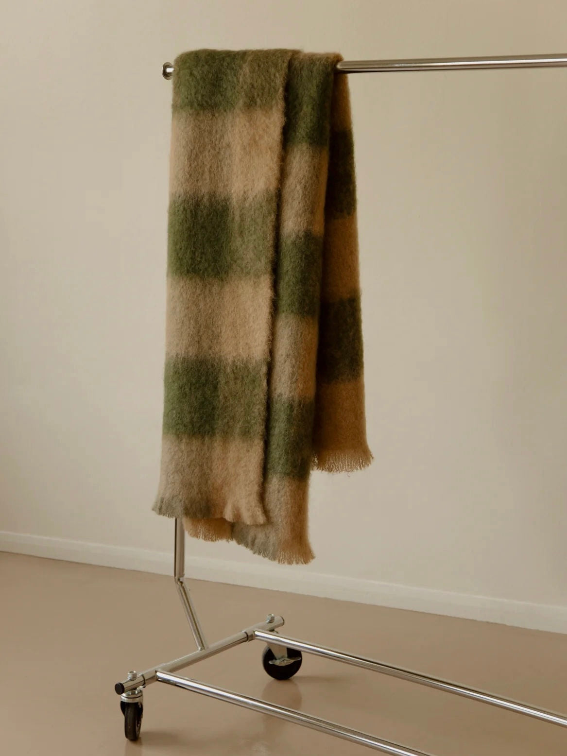 An elegant "Arbor no. 2" Throw by The Wren, featuring a large green and beige plaid pattern, drapes over a minimalist silver metal clothing rack in a softly lit room with beige walls and flooring.