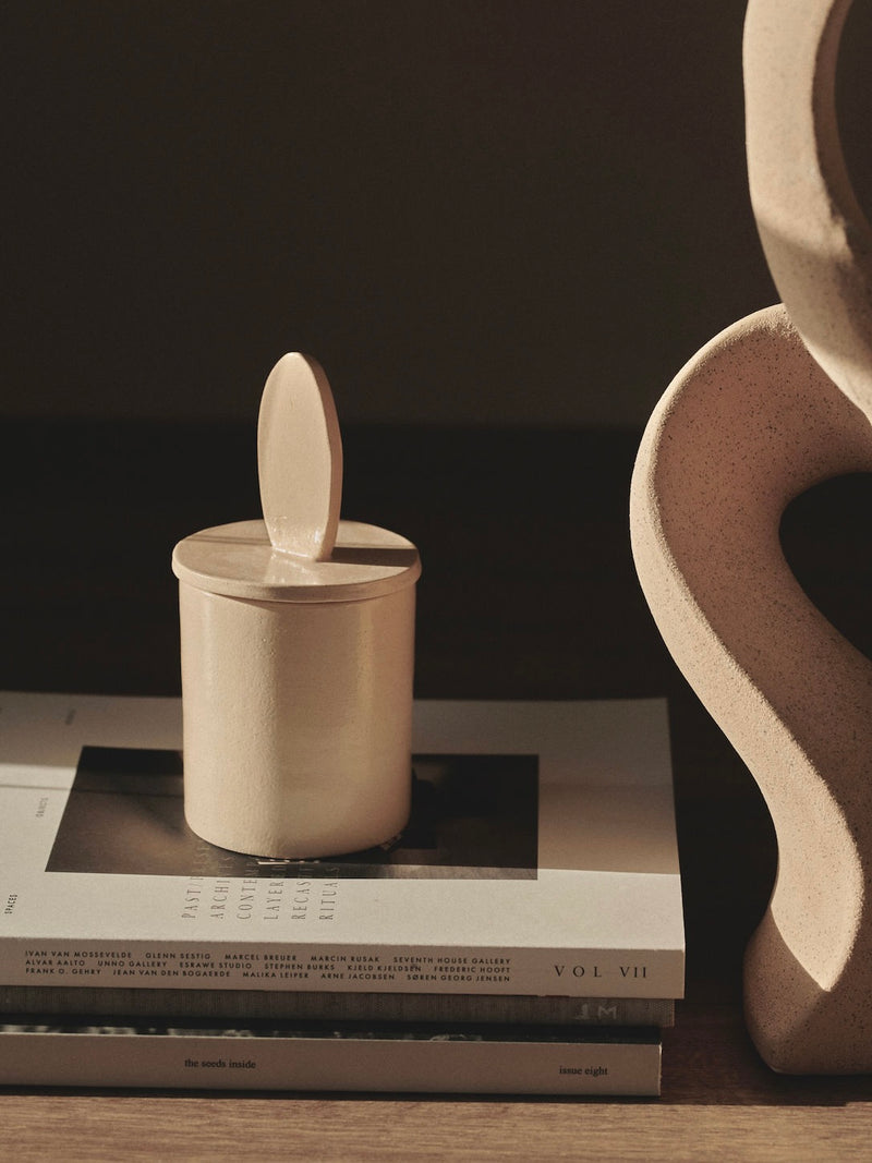 The Sculptural Scented Candle by 2222Studio, housed in a beige ceramic container with a handle, rests on stacked books under soft light. In the background, a curved textured sculpture is partially visible, with neutral tones and shadows creating a calm ambiance.