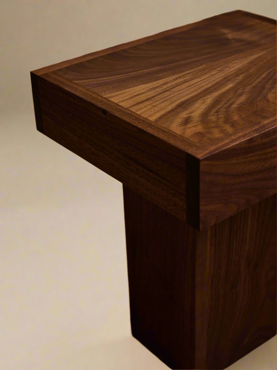 A close-up of the Jura Pedestal from Ah Um Design Studio, showcasing its natural, rich brown grain pattern. This handcrafted piece boasts a simple, modern design with clean lines and a rectangular top. The background is softly blurred, featuring a light-colored curtain and a portion of a carpeted floor—ideal display furniture.