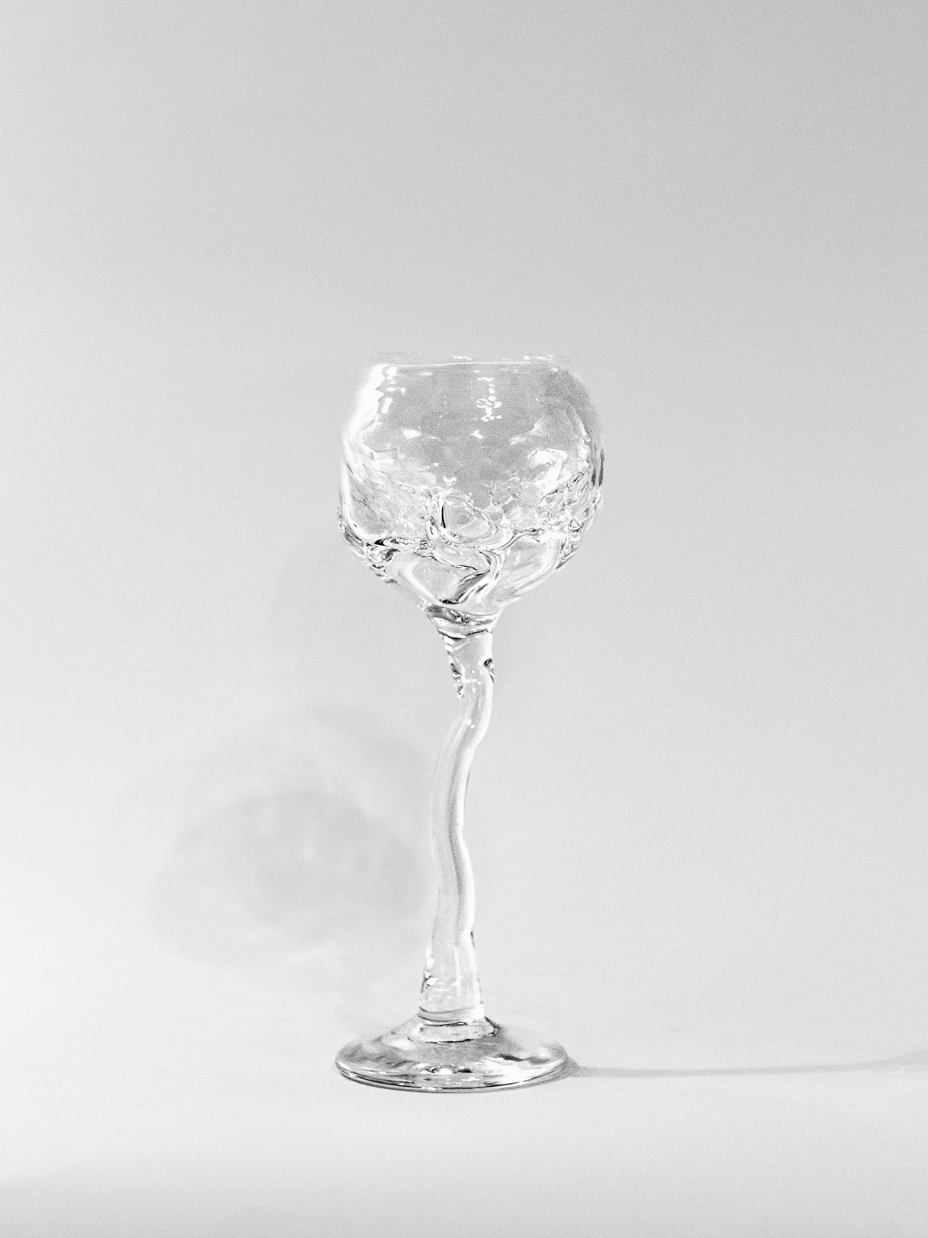 A clear Hand Blown Freeform Wine Glass by Szkło Studio, crafted from transparent sodium glass with an elegantly twisted and decorative textured bowl, sits on a plain white background, casting a faint shadow that highlights its intricate design and smooth curves.