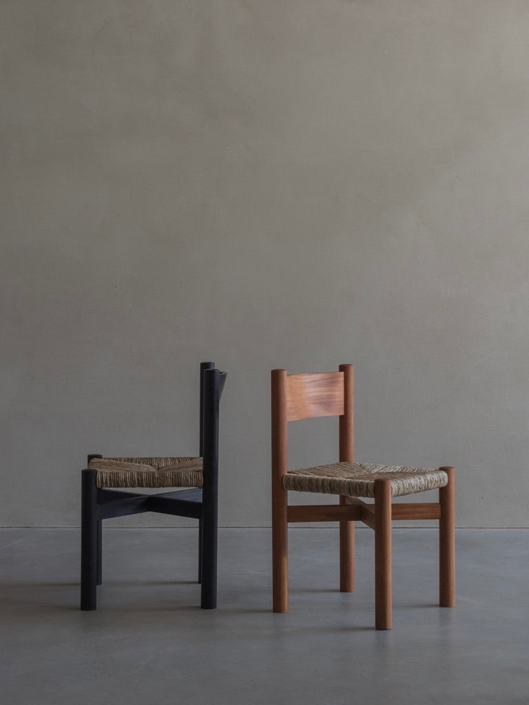 Two 'Les Allues' straw chairs by Bicci de' Medici are placed side by side on a smooth, gray floor against a plain wall. The left chair is black with a woven seat, while the right highlights mahogany craftsmanship. Both feature simple, straight backs and straw seats for rustic charm.