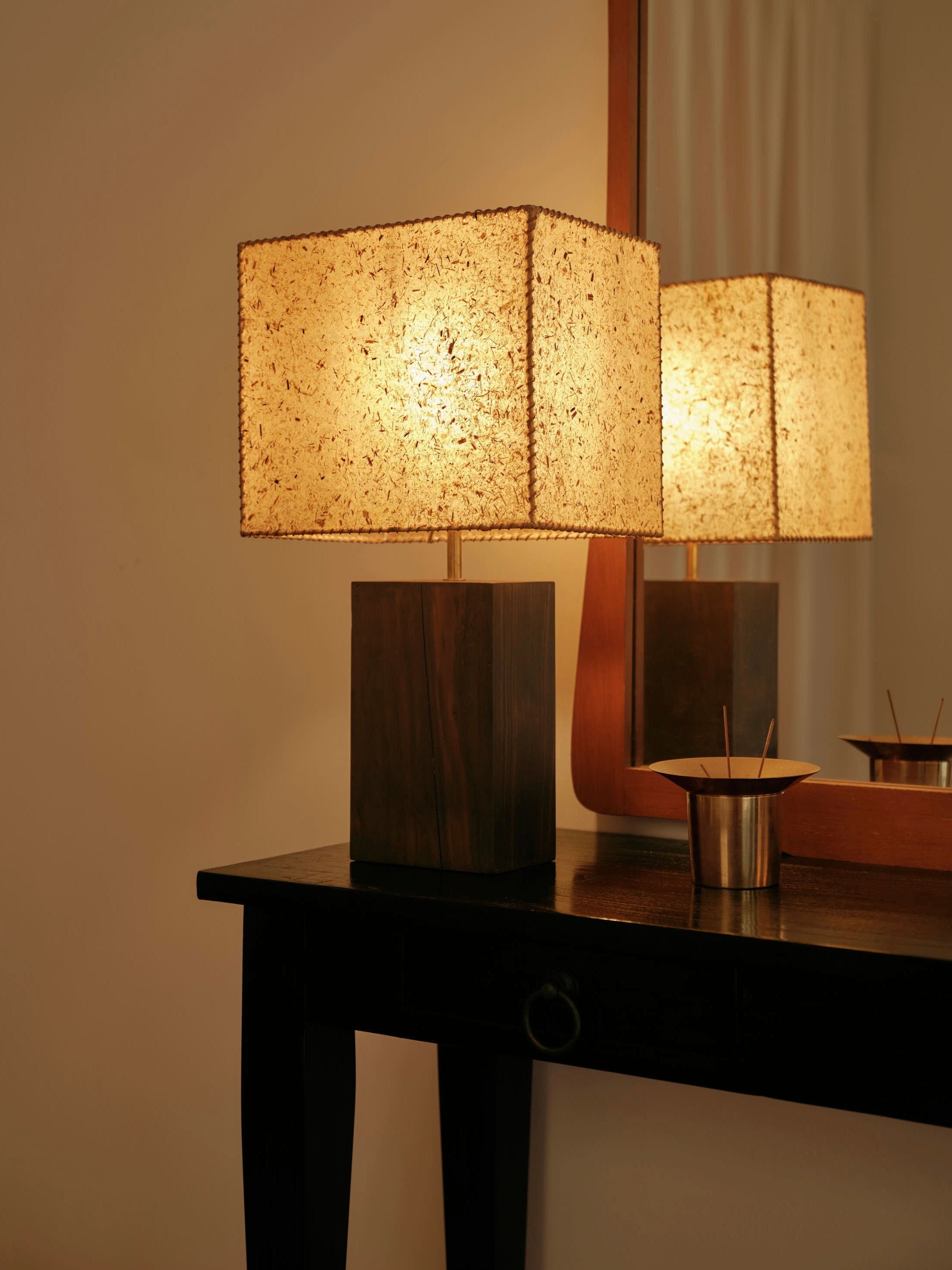 A warm, softly glowing 1948 Table Lamp by Lana Launay, featuring a textured beeswax shade, sits on a dark wooden console next to a small metallic container. A mirror behind reflects the lamp's light, adding a cozy ambiance to the room.