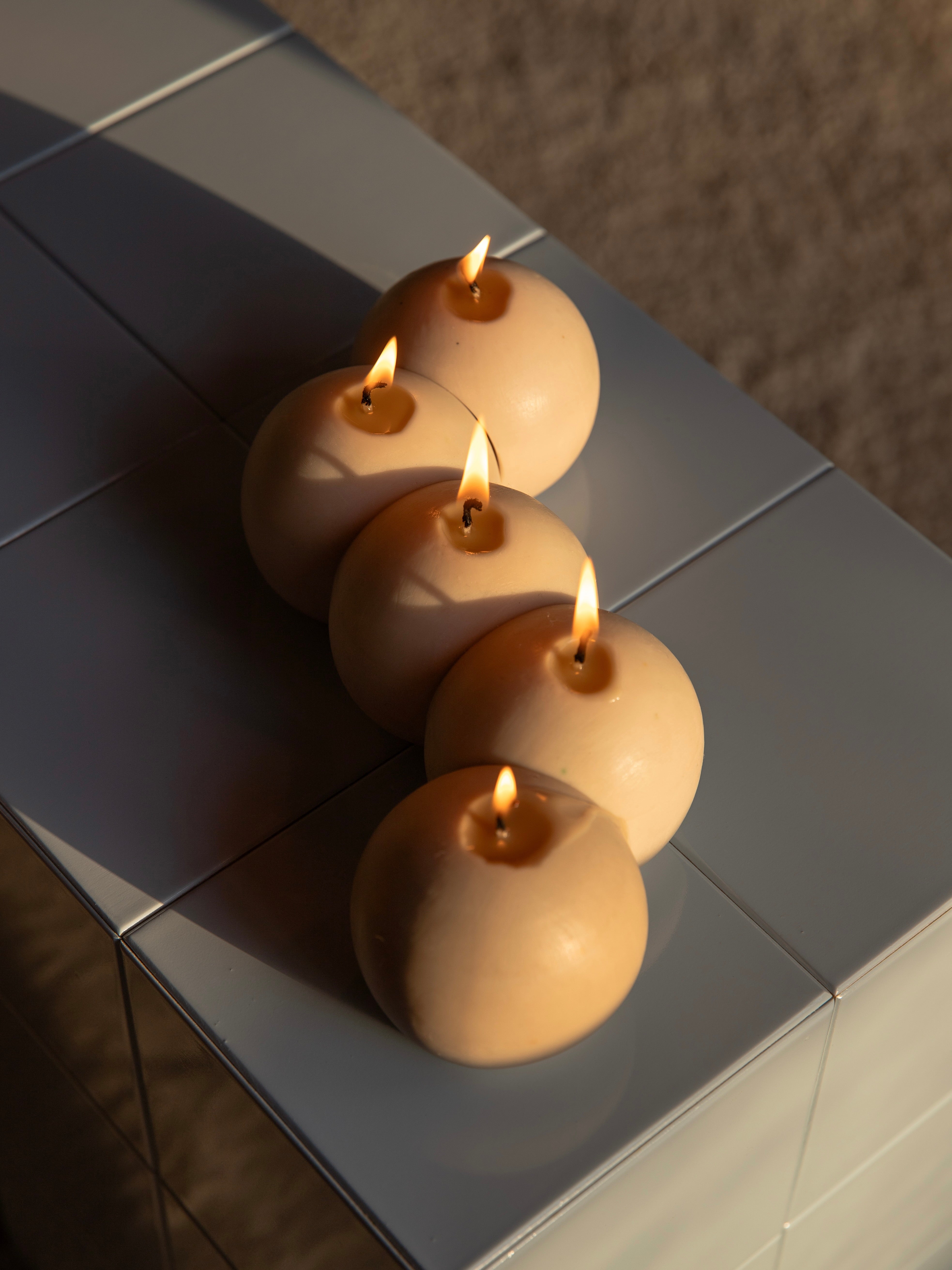 Five E15 candles from Garado Studios are lined up on a shiny surface, their small flames casting warm light that contrasts with the cool-toned background, evoking an Olafur Eliasson installation and creating a cozy, serene atmosphere.