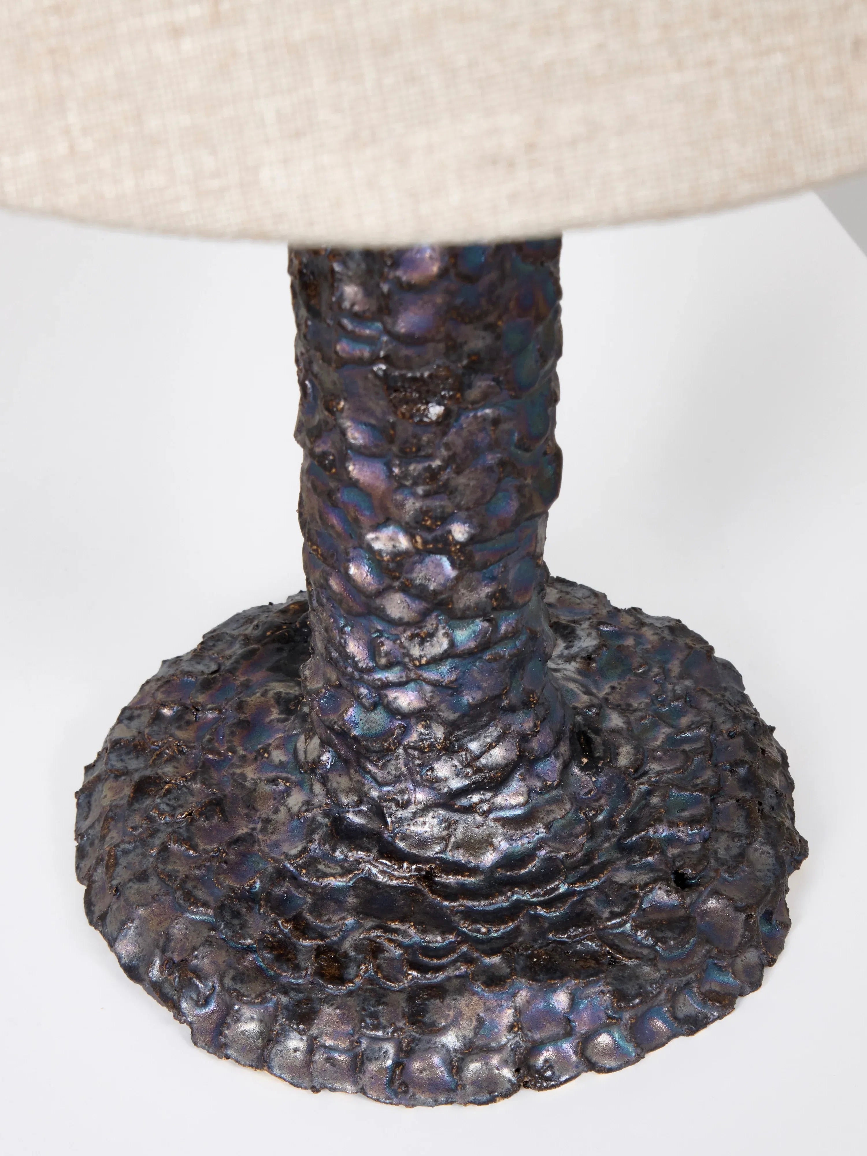 Close-up of the Casa Bedside Light by Project 213A, featuring an artisanal ceramic base with a reptile-like iridescent scaled texture. The dark base shimmers in purple and blue, partially covered by a neutral textured lampshade.