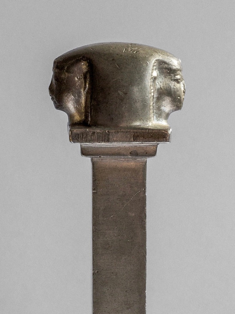 A metallic object with a rectangular stem extends upwards to a top that features two sculpted heads facing in opposite directions, resembling an Art Deco Seal Stamp Sculpture from the 1930s by Spigel, Sweden. The heads appear to be of ancient figures with intricate headgear, and the entire object has a worn, vintage appearance.