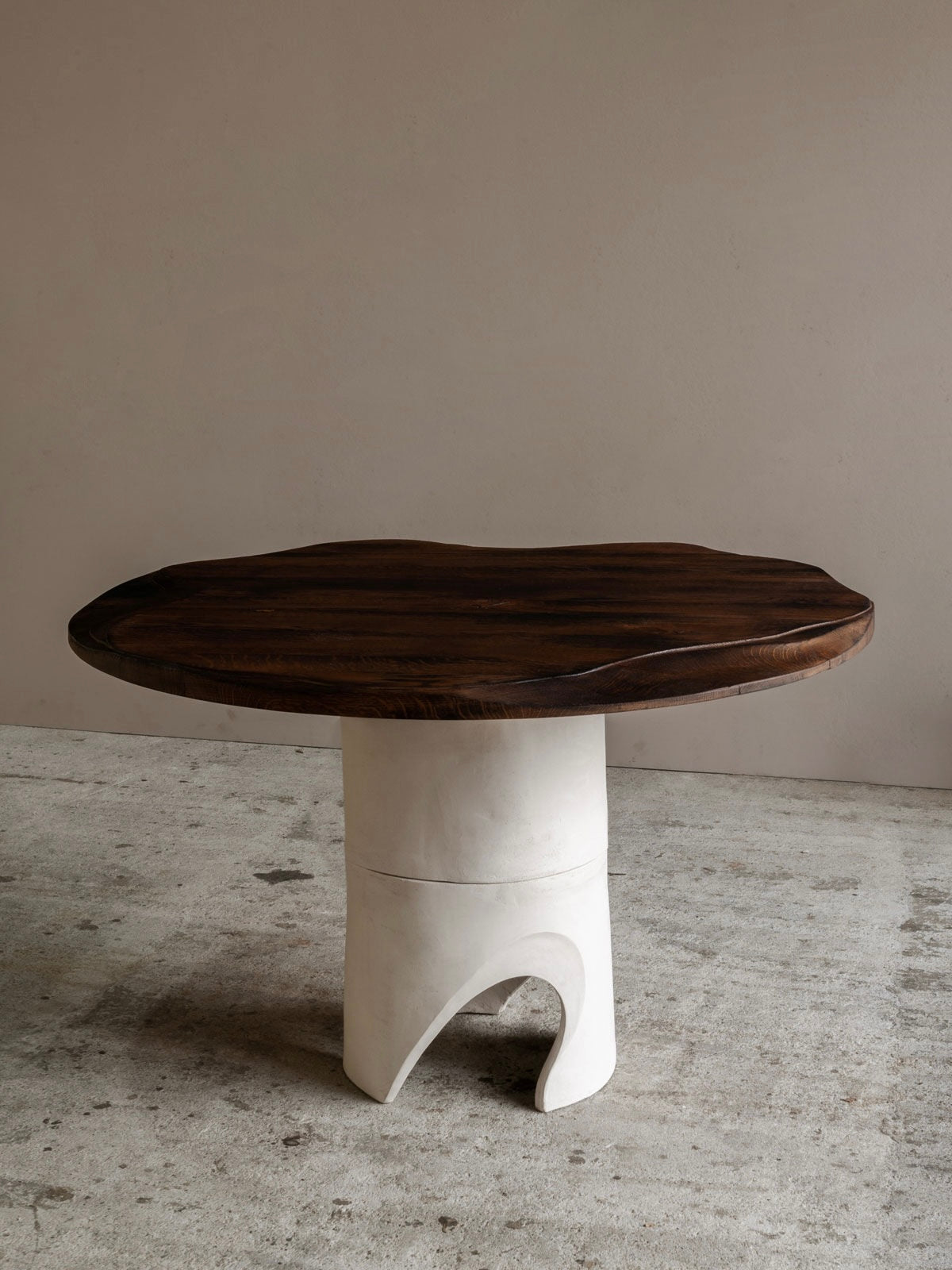 The Lac - Carved Table in Sandstone and Oak by Lea Bigot features a round wooden top with a dark, irregular finish set on a sculpted white base with an arched opening, showcasing modern design. It sits on textured concrete against a plain beige wall.