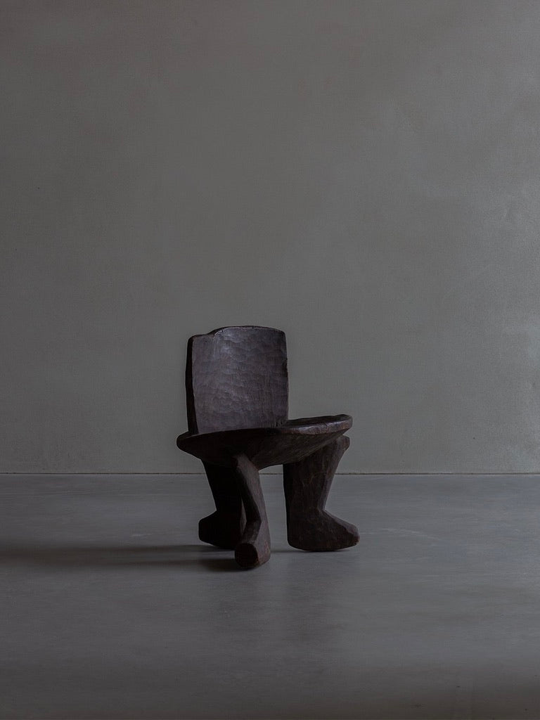 A Bicci de' Medici African Stool with a dark, sculptural wooden design stands on a gray concrete floor against a plain gray wall. Featuring an abstract, rough texture and asymmetrical legs, it reflects the skill of African artisans in preserving cultural heritage.