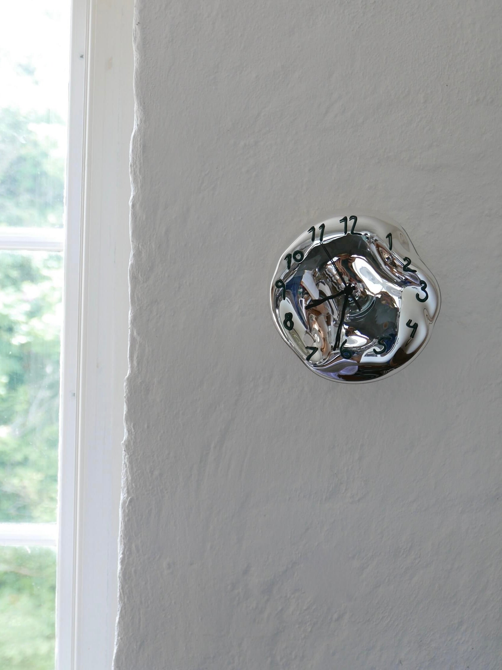 The Silverclock - Green and Black, a handcrafted wall clock by Silje Lindrup from Denmark, is mounted on a light gray wall adjacent to a window on the left side. This silver clock features a face that appears to be melting or warped, with twisted numbers and hands distorting their usual positions. Through the window, there is a glimpse of greenery outside.