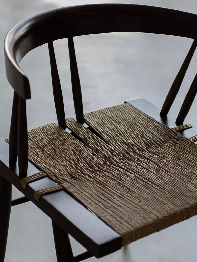 The "Manner of Nakashima - Grass Seated Chair" by Bicci de' Medici is a stylish wooden chair with a curved backrest and woven seat in neutral tones, exuding mid-century allure. Its elegant design features clean lines with a dark rosewood finish on a subtle gray background.