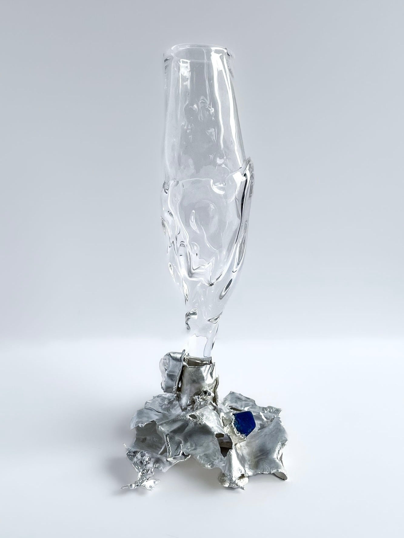 The Jewelled Goblet no. 10 by Lindsey Fontijn, from Szkło Studio, showcases a modern glass sculpture with an elongated abstract hand-blown glass form mounted on a jagged metallic base. The base is adorned with a small blue accent, and the sculpture's sleek and artistic design evokes the elegance of silver-stemmed goblets, set against a plain white background.