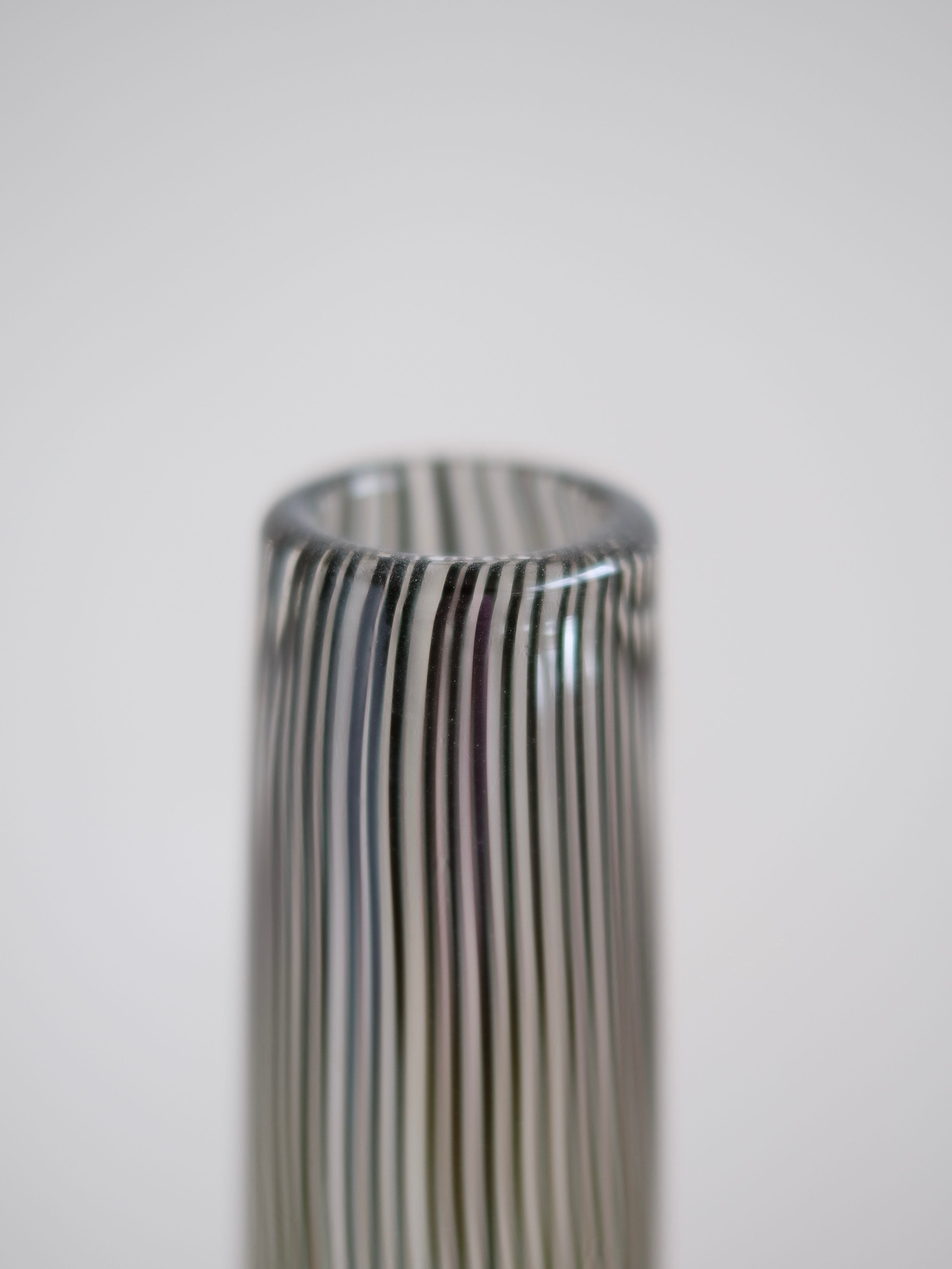 Close-up of a cylindrical Italian Murano Vase, 1970 by septembre studios, featuring vertical black and white stripes that create an optical illusion effect. The surface looks smooth and glossy, with a slightly rounded top edge. The background is softly blurred in a neutral tone.