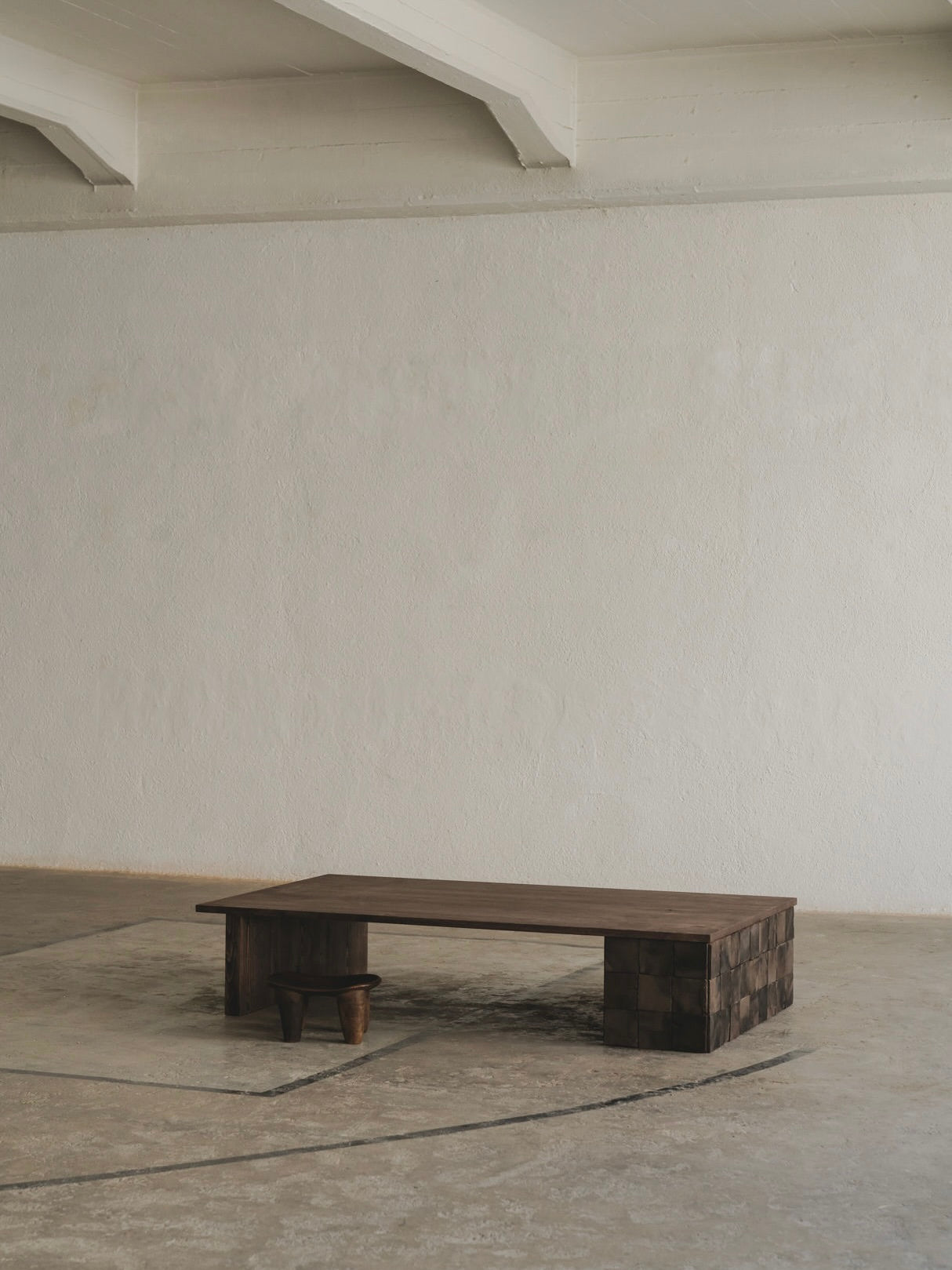 In a minimalist room with a plain beige wall and concrete floor, a long European oak bench with a unique finish sits centrally, accompanied by a small round wooden stool reminiscent of the Cosmos Low Table by ASERIES OF OBSTACLES, SL. The space is empty yet well-lit.