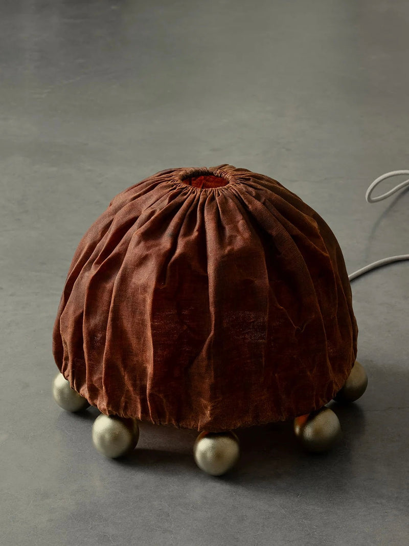 The ALTER Table Lamp by Kaytar features a dome-shaped lampshade made of beeswax-coated linen with a gathered top, supported by spherical brass legs on a gray surface. A cord extends from the back, highlighting its handcrafted, artisanal design.