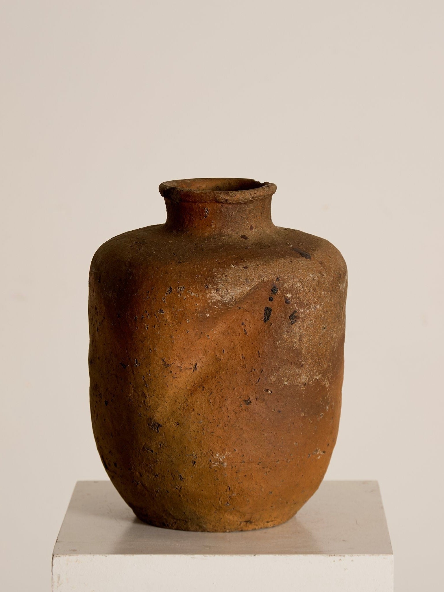 The Japanese Stoneware Vessel by Ancien et Jolie, featuring a rustic terracotta finish with speckle glaze and dark speckles, exudes antique Kasam Japan charm on a white pedestal against a light background.