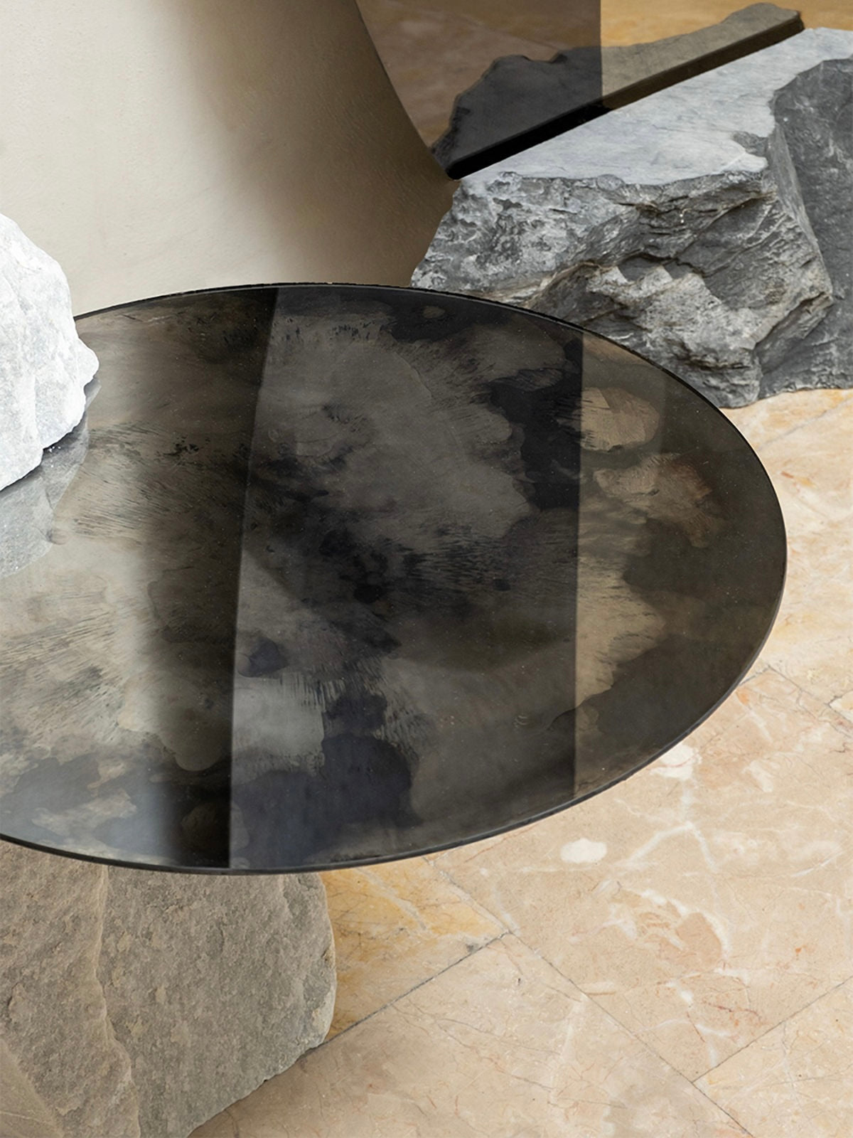 The Cloud Mirror Side Table by HOMA features a round glass design with dark, cloudy patterns on rough stone slabs. Its background includes oxidized silver mirrors and stone textures, creating an earthy, minimalist aesthetic where light and shadows enhance its unique elegance.