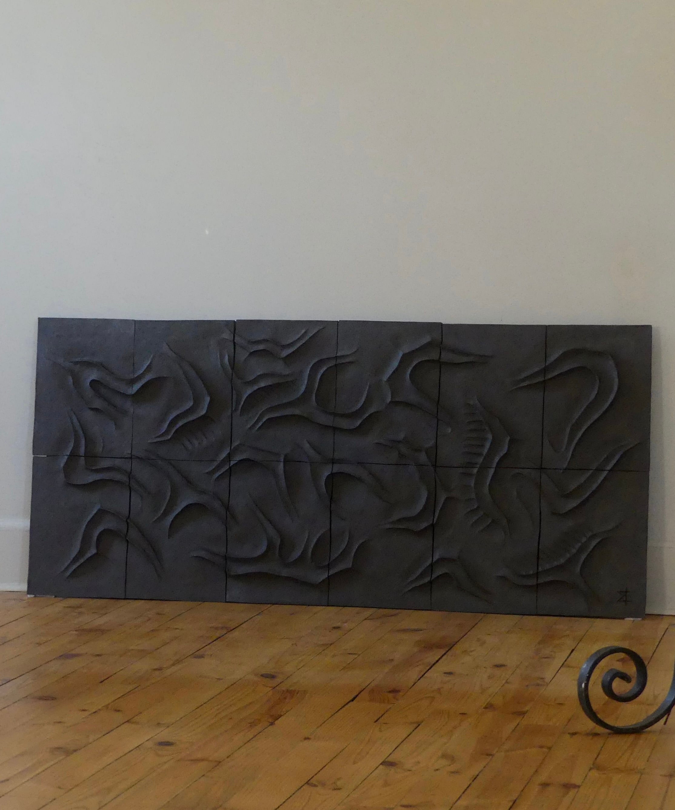 The Caldeira Wall Sculpture by Franck Scala is a dark, abstract ceramic bas-relief composed of curved square panels, set against a light wall on a wooden floor. A metal element in the lower right hints at its inclusion in the RELIEFS 2025 Collection.