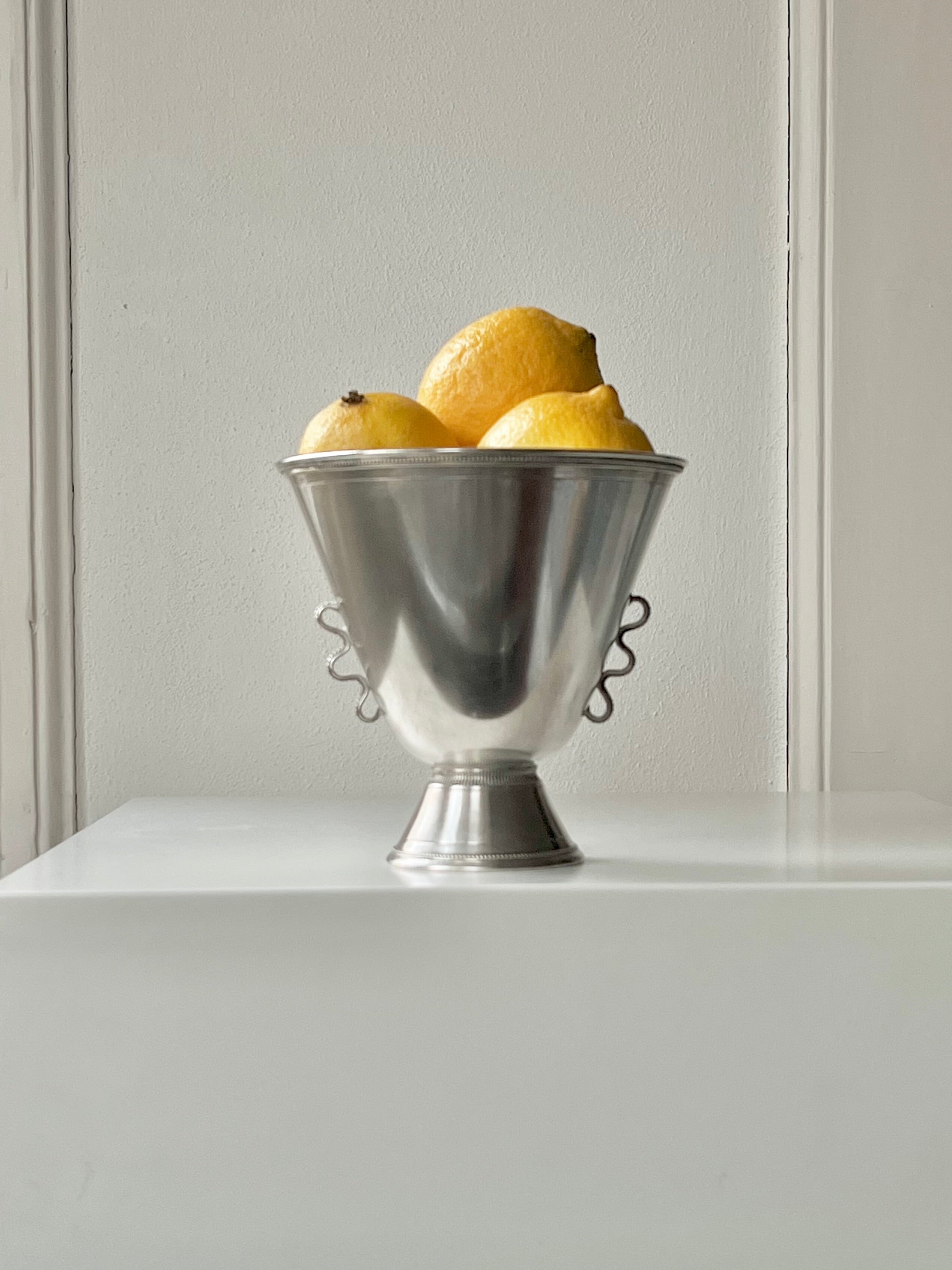 A 1930s Médecine silver vase holds four bright yellow lemons, elegantly placed on a white surface against a neutral background. This minimalist and fresh composition echoes the understated elegance of a Stockholm design studio.