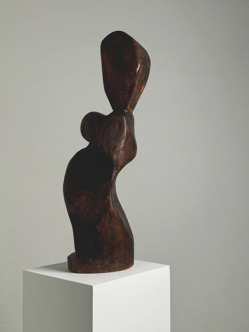 The Large Anthropomorphic Wood Sculpture by Figuresdesign, featuring exaggerated curves, smooth contours, and an organic form reminiscent of a woman's silhouette, is displayed on a white pedestal against a plain background, evoking movement and fluidity.