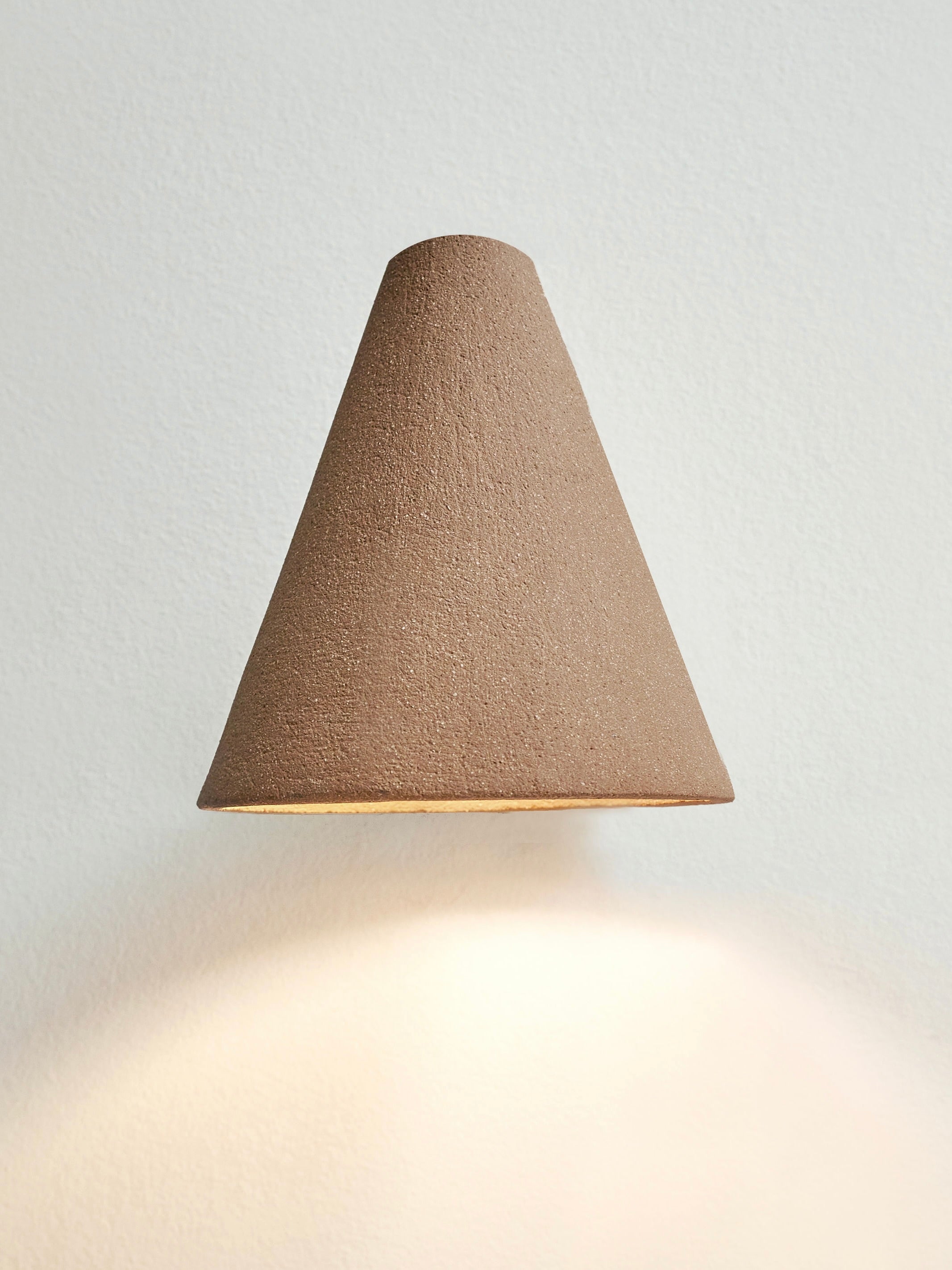 The 'Esoteric' Almond Brown Wall Light by Mariza Galani is a handcrafted, wall-mounted lamp with a cone shape and textured surface that mimics stoneware. It emits a soft glow onto light walls, offering an artisanal touch to any space.