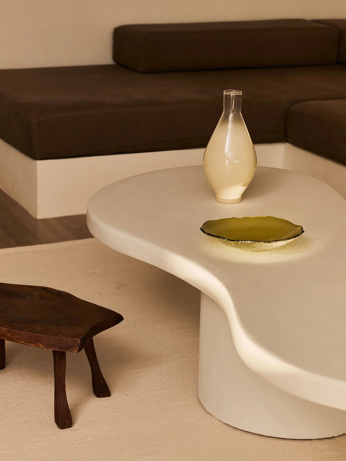 A modern living room showcases the uniquely shaped "Pesto" Coffee Table by MarlotBaus, crafted from artisan microcement. The table holds a pale yellow vase and a round green glass dish. Nearby, there is a small wooden stool, while a brown sectional couch lines the wall on the carpeted floor.