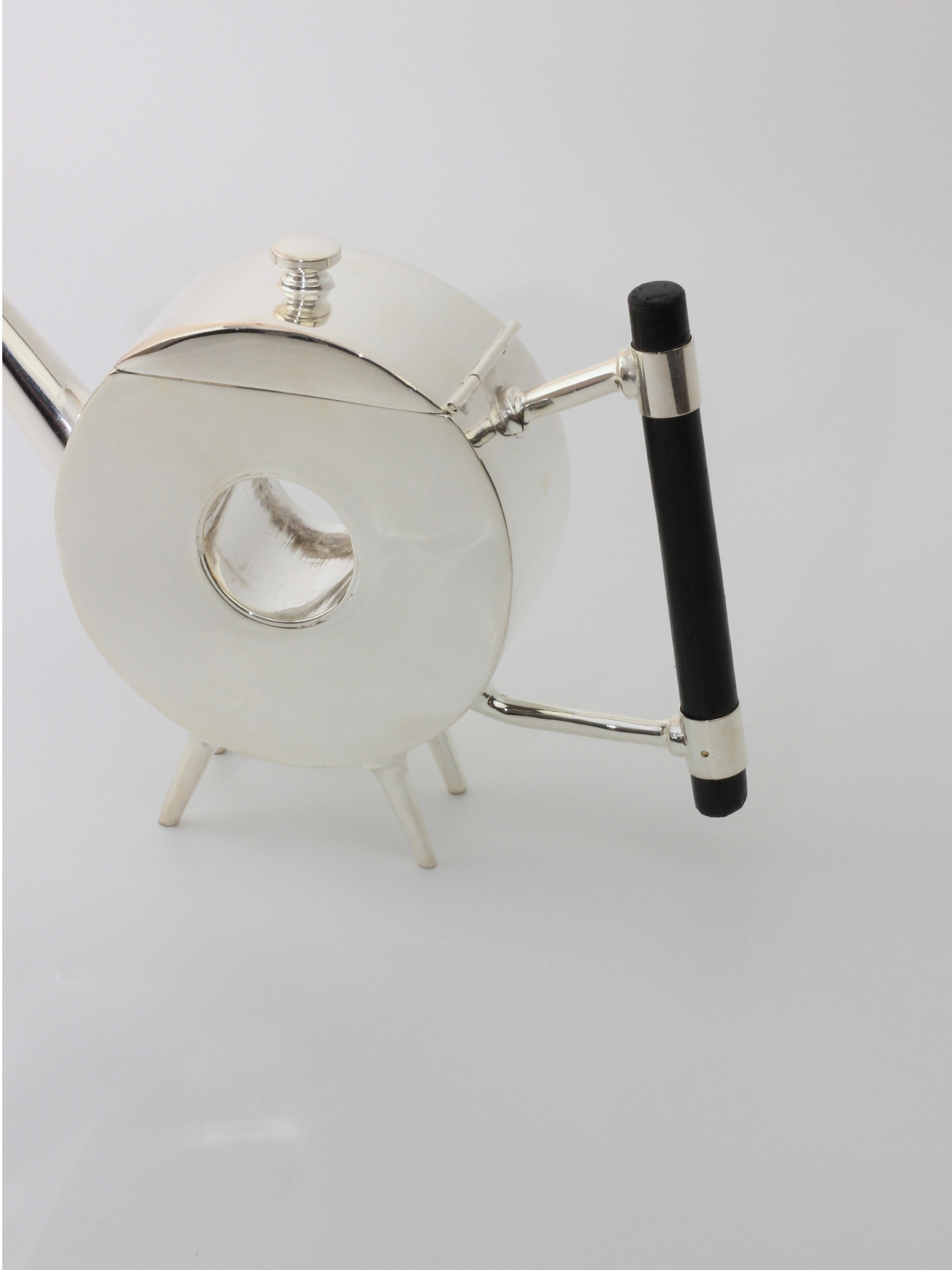 Christopher Dresser's Tea Pot from Boga Avante Shop, a modern, silver-plated teapot with a circular body and three short legs, sits against a plain background. It features an ebonized wood handle attached at a right angle and has a small round lid on top. The narrow spout extends straight out from the body, echoing Dr. Christopher Dresser's iconic designs.