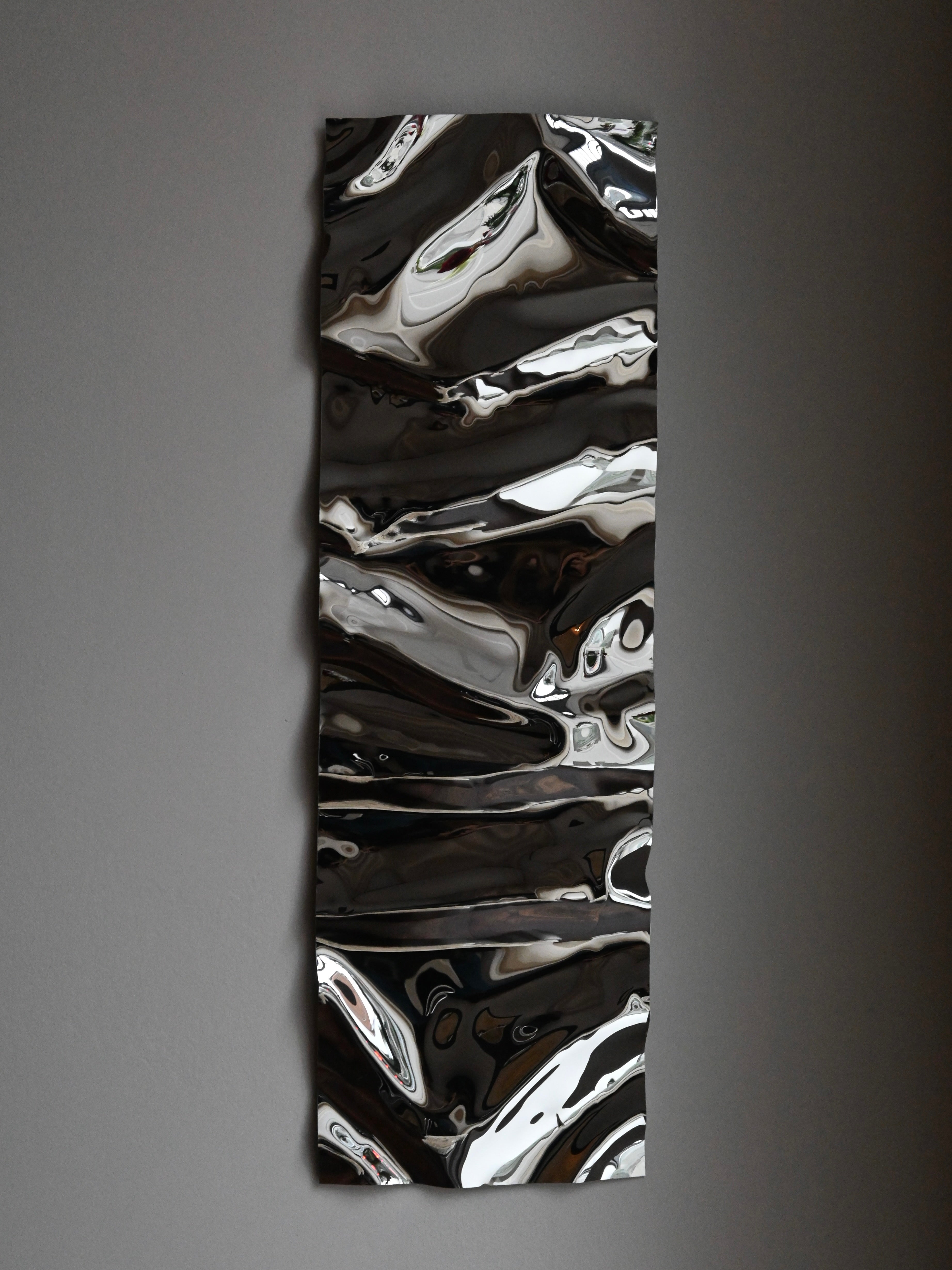 The "Psychedelic Mirror Long" by Caia Leifsdotter is a handmade abstract metallic wall sculpture with a wavy design. Its reflective silver and gray surface creates a distorted mirror effect, enhancing any plain gray wall when hung vertically.