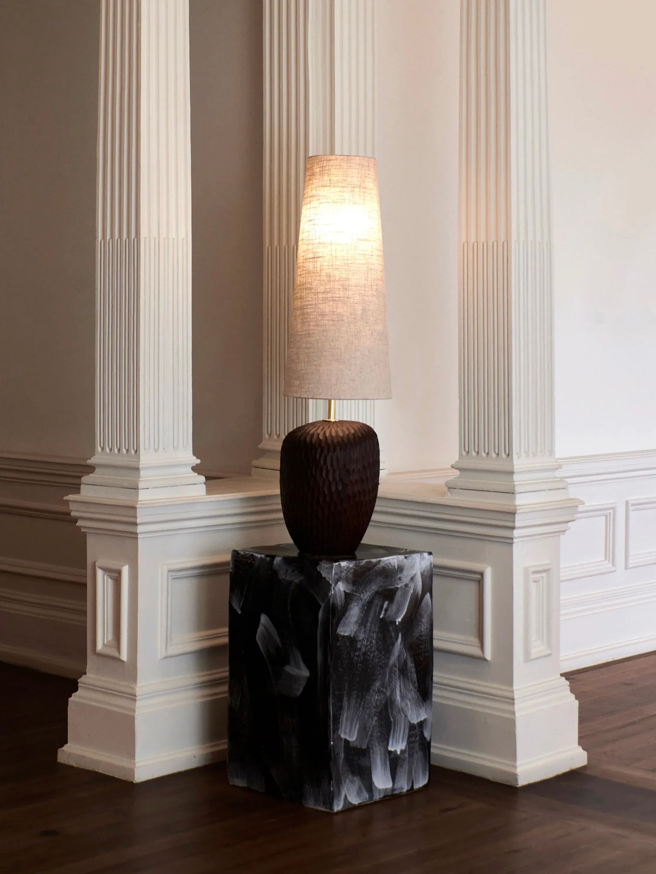 A tall fabric lampshade atop a textured dark brown base is placed on the Ceramic Side Table with a geometric shape from Project 213A. This setup is positioned between two white, ornately carved pillars in a minimally decorated, elegant room with wooden floors, adding modern flair.