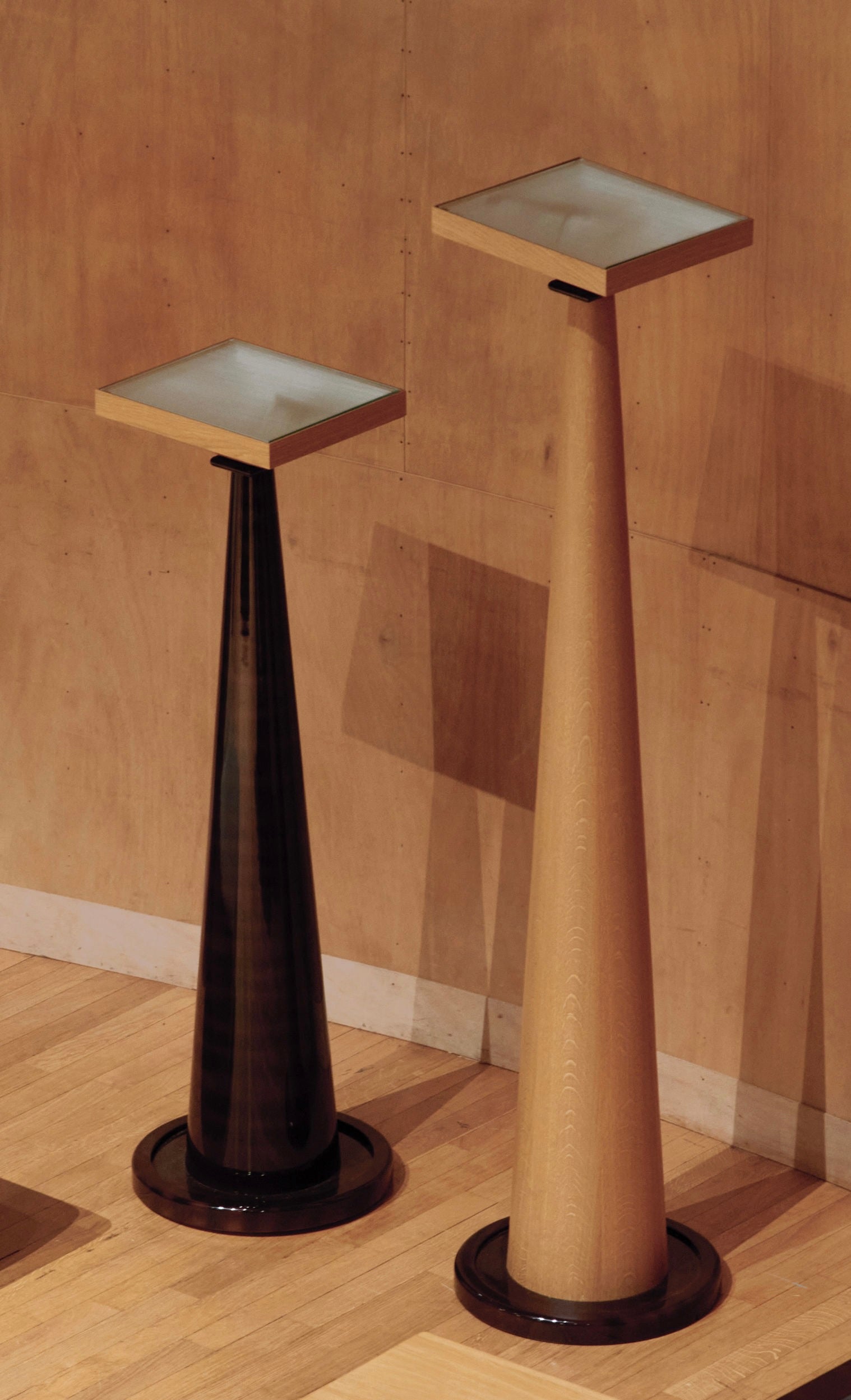 Two tall, conical "Le Fontainebleau" floor lamps by HAUVETTE & MADANI with square tops stand elegantly. One lamp features a darker shade with a glossy lacquered finish, while the other showcases a lighter oak hue with a matte finish. They are set against a wooden panel background on a light wooden floor.