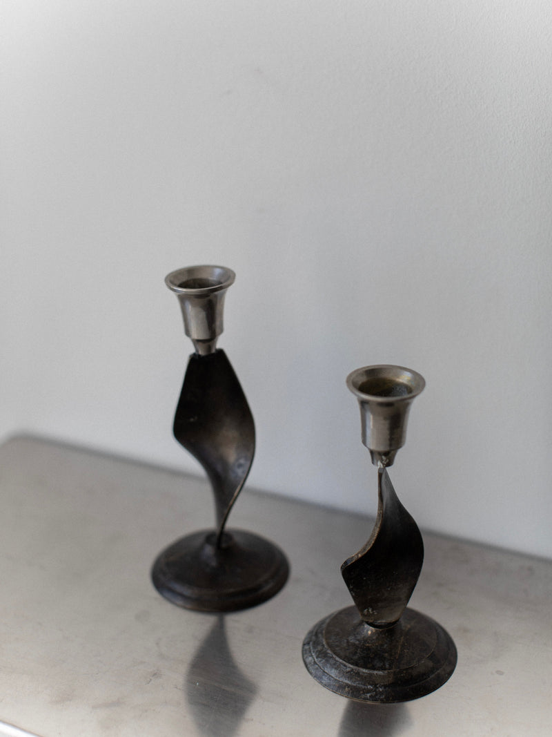 The Brutalist Candleholders by Out For Lunch, featuring a modern design in twisted black steel, stand on a reflective surface against a light-colored background, adding a sophisticated touch reminiscent of the 70's to your home decor.